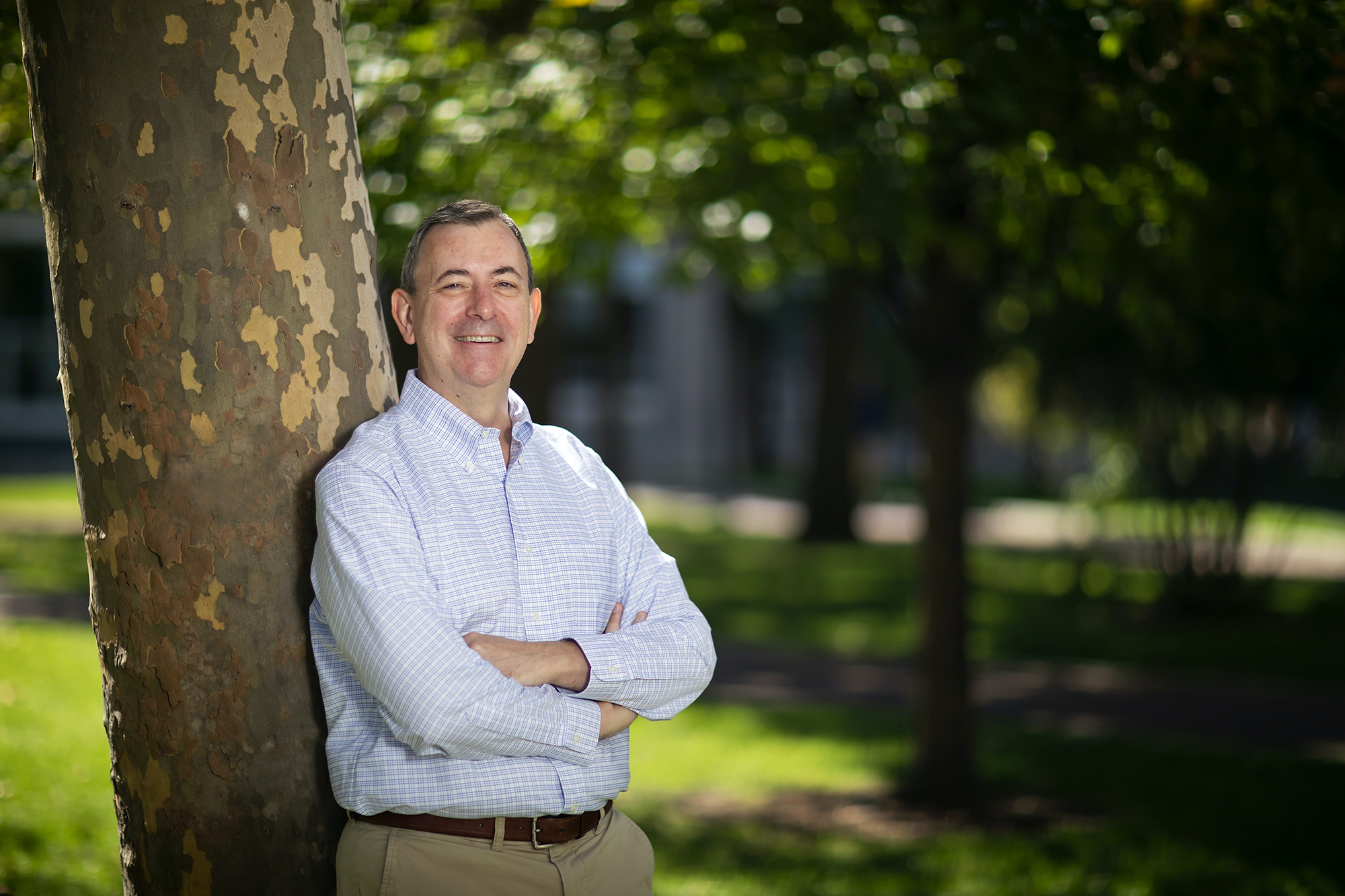 A person leaning against a tree outside, wearing a blue-and-white button down shirt, arms crossed.