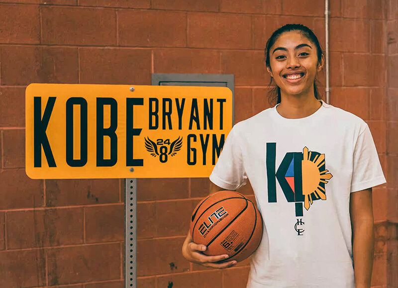 Kayla Padilla smiles while holding a basketball in a t-shirt she designed in front of a sign that reads Kobe Bryant Gym.