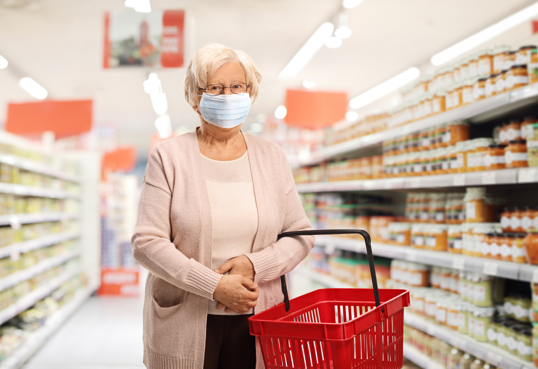 Senior citizen wearing a mask holding a shopping basket in a grocery store aisle.