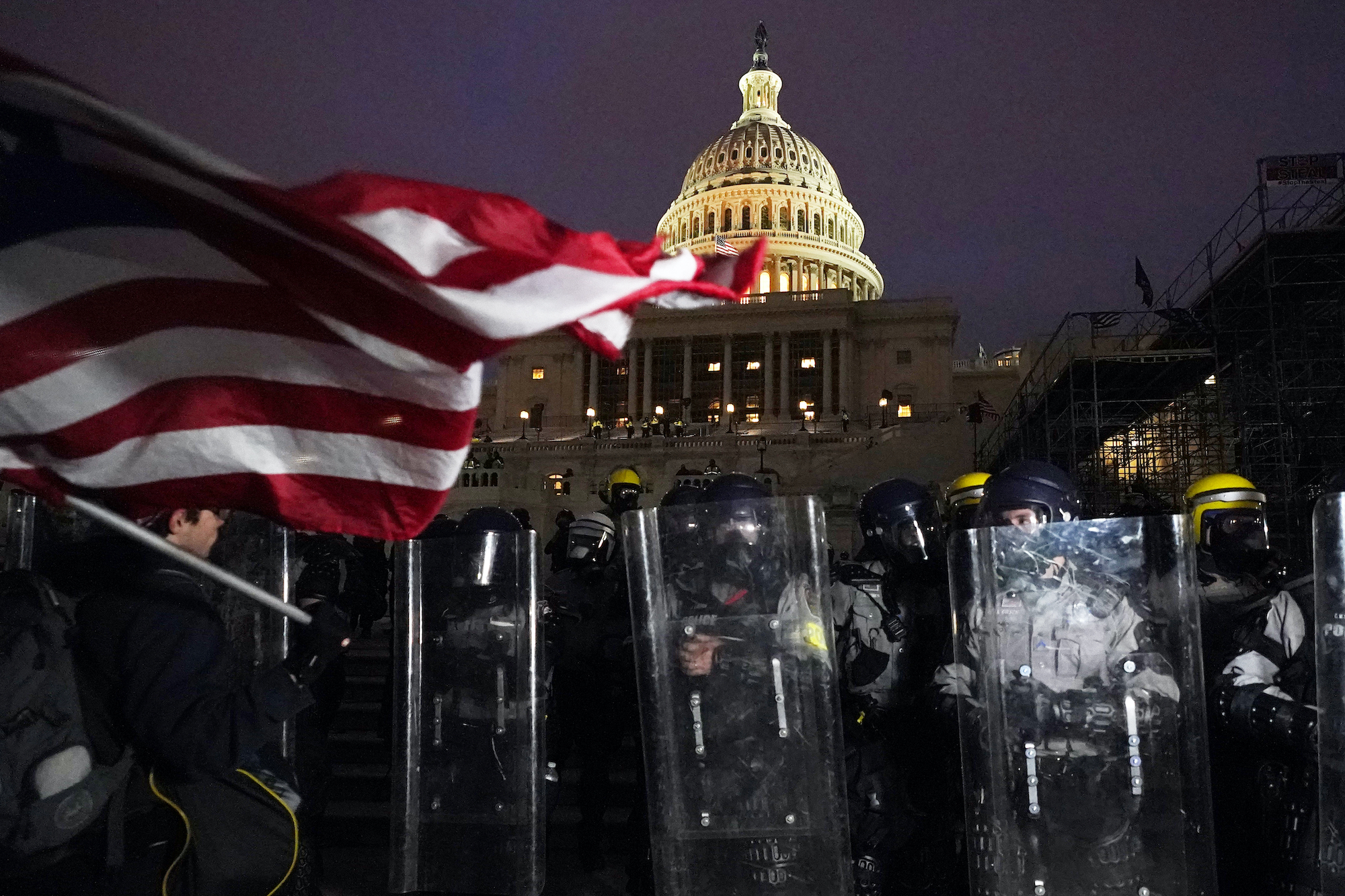 Riot police hold their shields in front of the U.S. Capitol, lit up at night, as a man with an American flag looks on