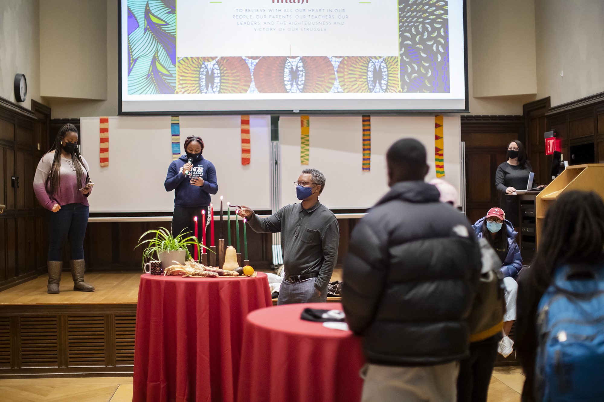Students watch a man lighting seven red and green candles for Kwanzaa. The table also displays plants, squash, and maize in representation of the harvest festival