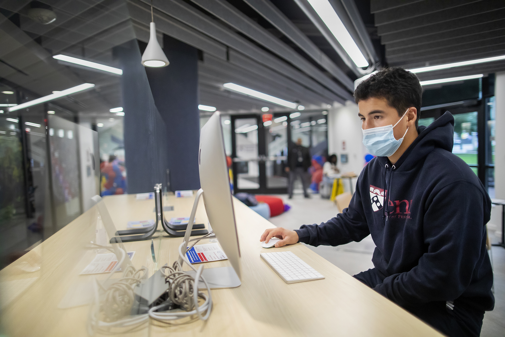 A man wearing a mask and a Penn hoodie uses a desktop computer at a high top desk in a lobby