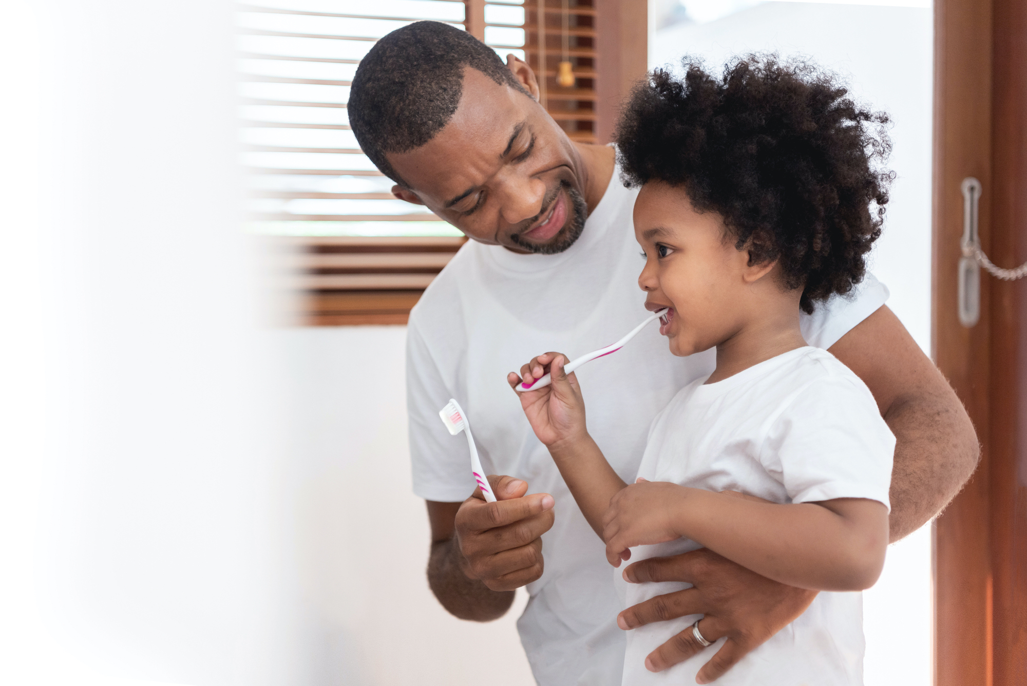 Adult holding a toothbrush standing with arm around toddler brushing teeth.