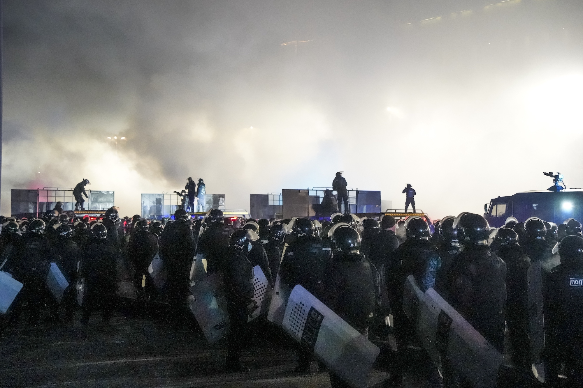 Protesters and riot police stand on a street in Almaty, Kazakhstan, as smoke rises in the background