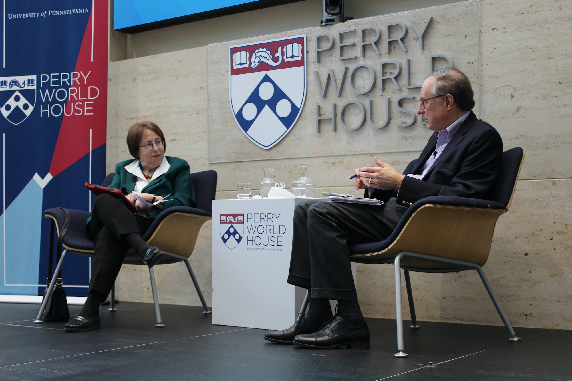 Trudy Rubin and former U.S. Ambassador to Russia Alexander Vershbow sit in chairs on a stage with a sign between them reading Perry World House
