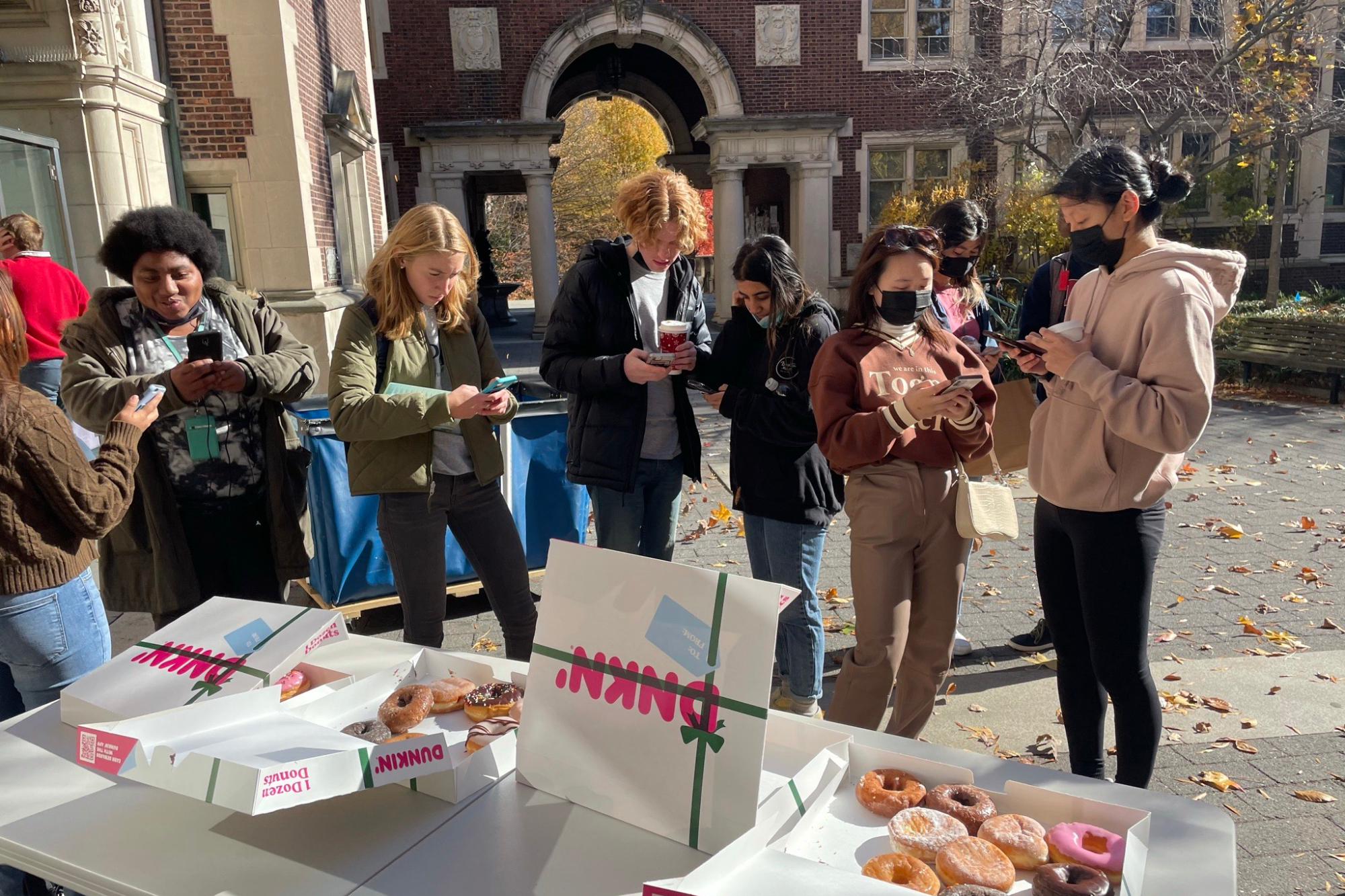 people outside wearing masks taking surveys on their phones while standing next to a table covered with donuts