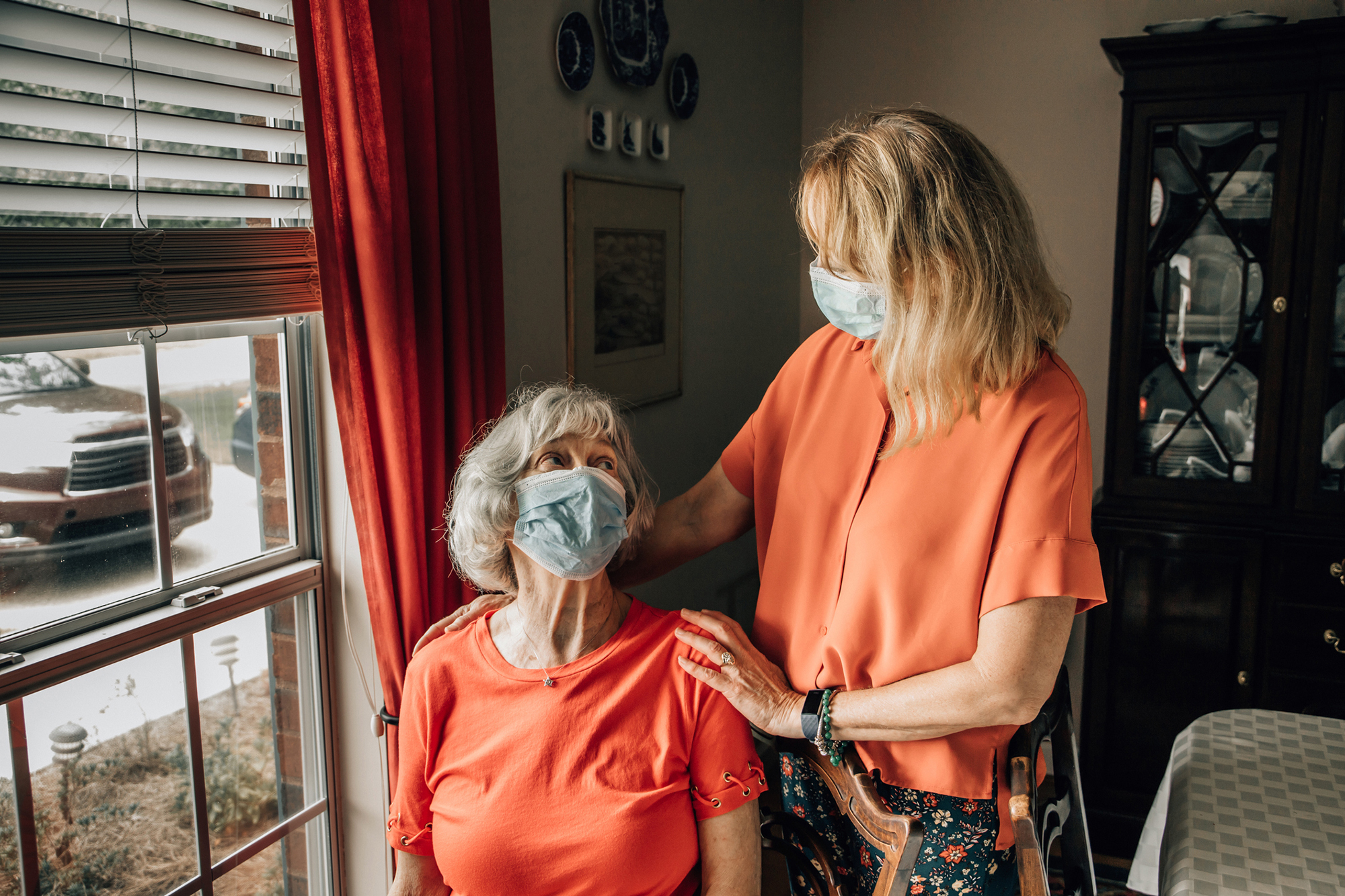 Elderly person wearing face mask in their home at a window being cared for by an adult also wearing a face mask.