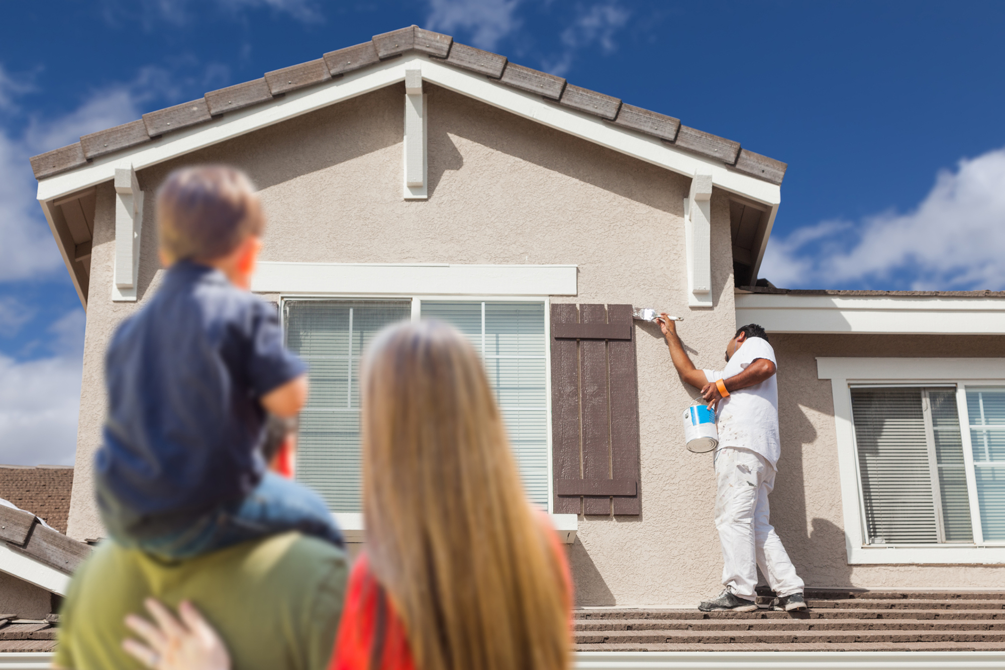 Two adults and a child watch a housepainter painting shutters on the front of the house.