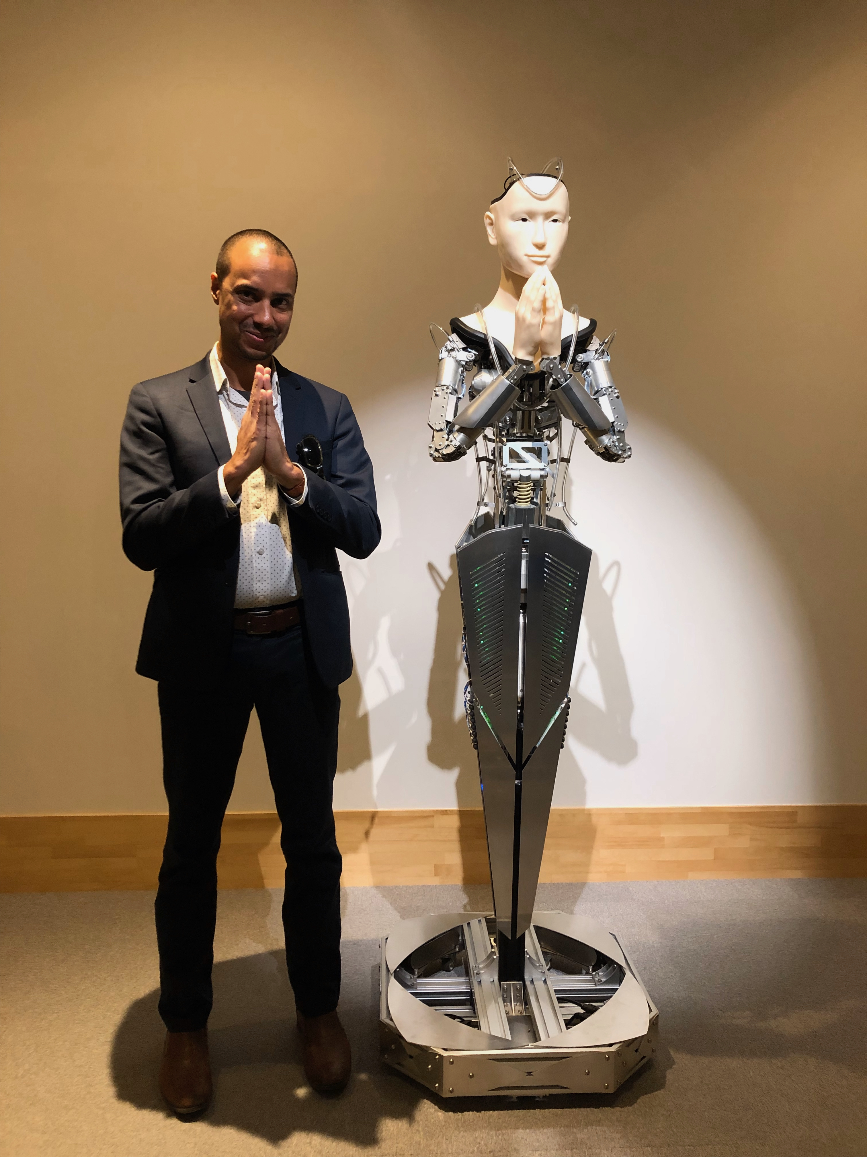 Man with prayer hands stands next to an android figure in a similar pose