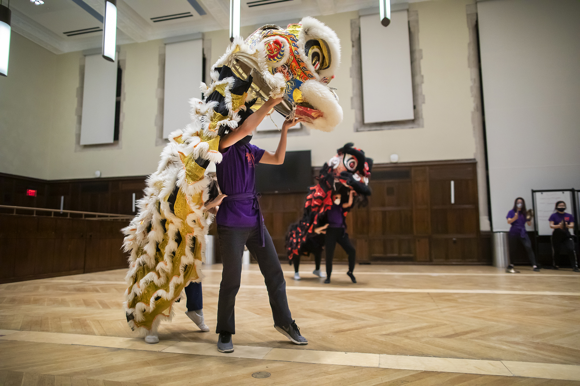 Two pairs of lion dancers practice in a large auditorium with cymbal players in the background