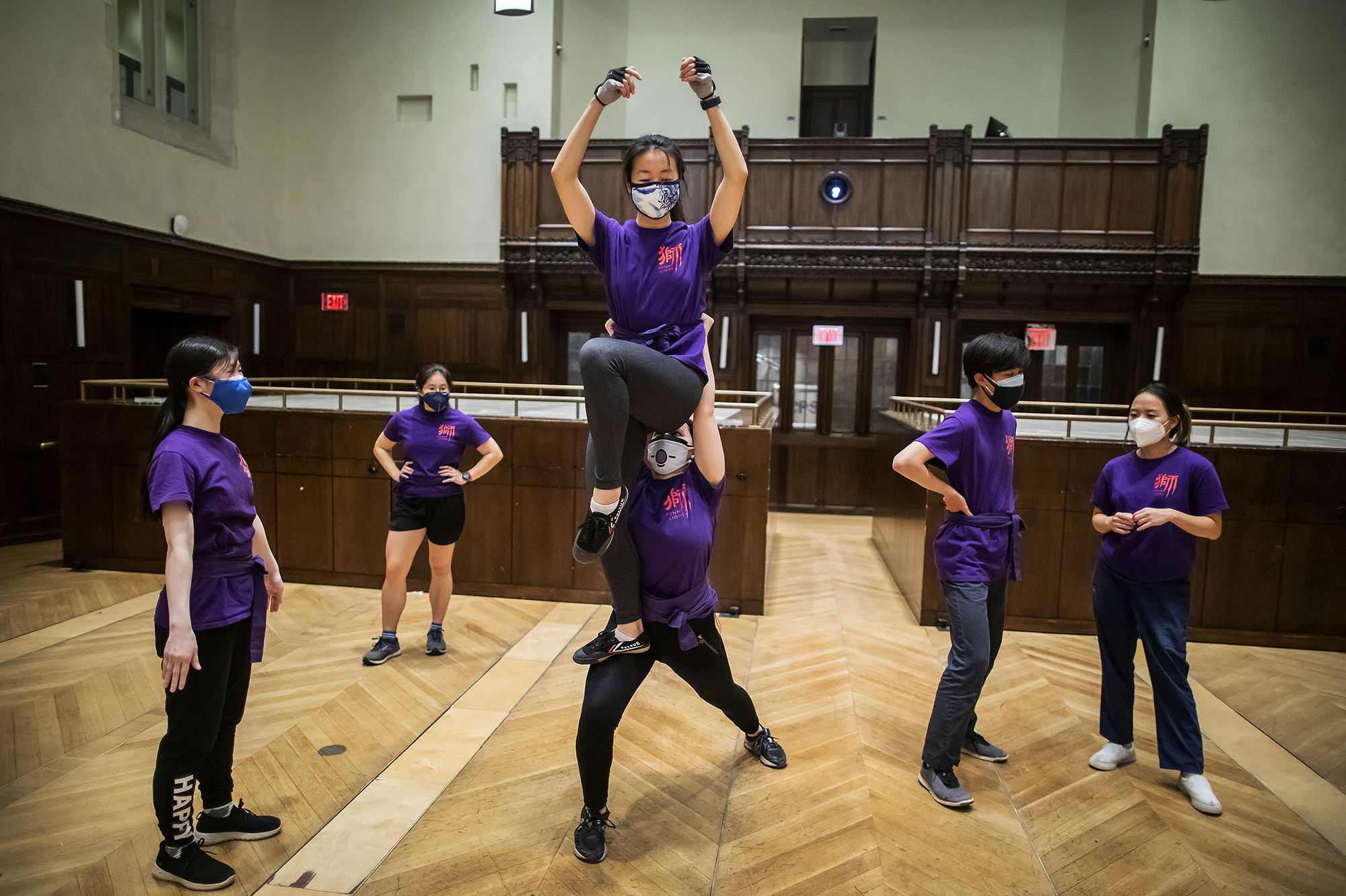 Three pairs of dancers are in identical purple shirts; the middle pair is practicing a "stack," with one dancer perched on top of another's head. The raised dancer has her arms up, pretending she is holding the lion head.