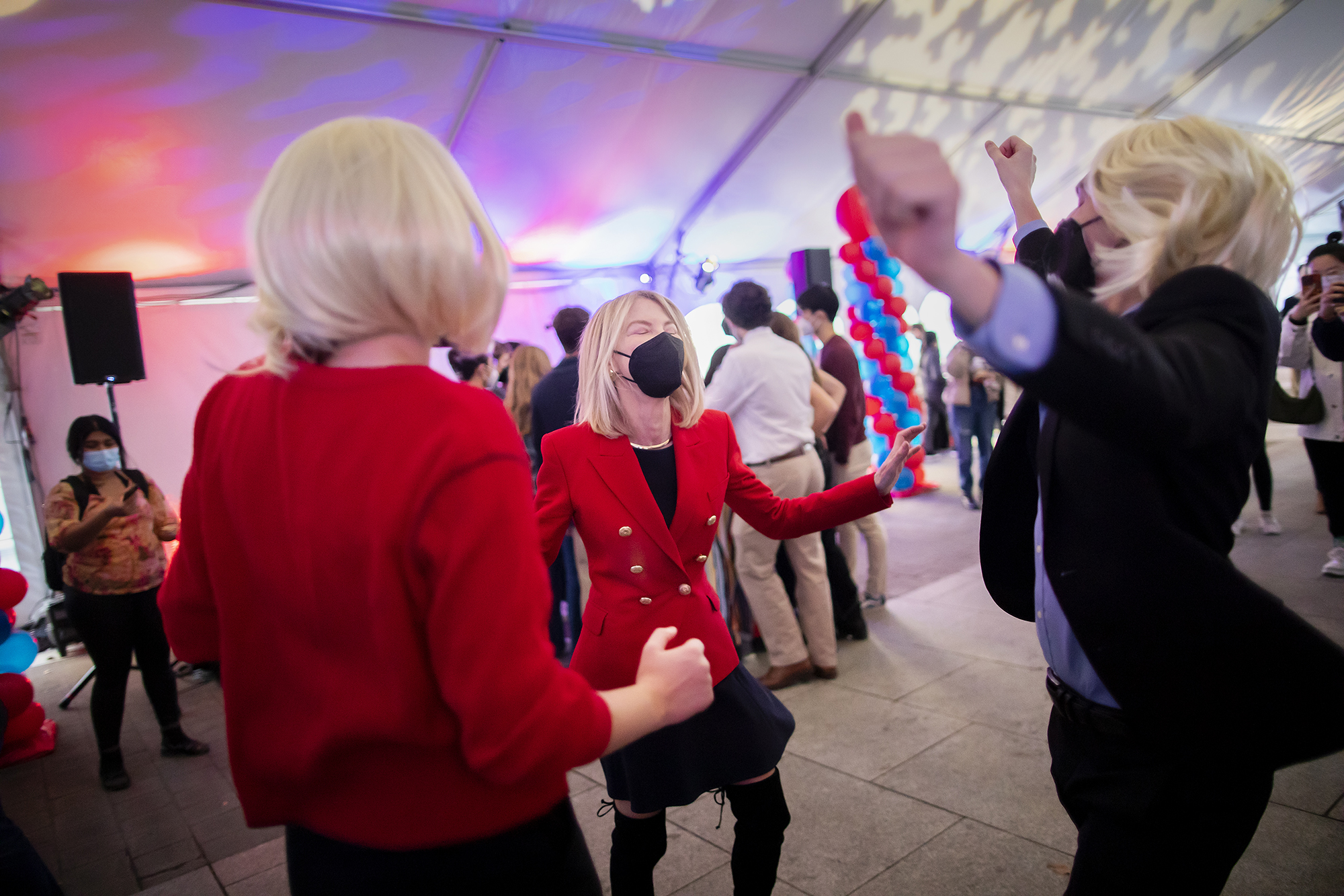Penn President Amy Gutmann at a party talking with two students wearing blonde wigs with their arms in the air.