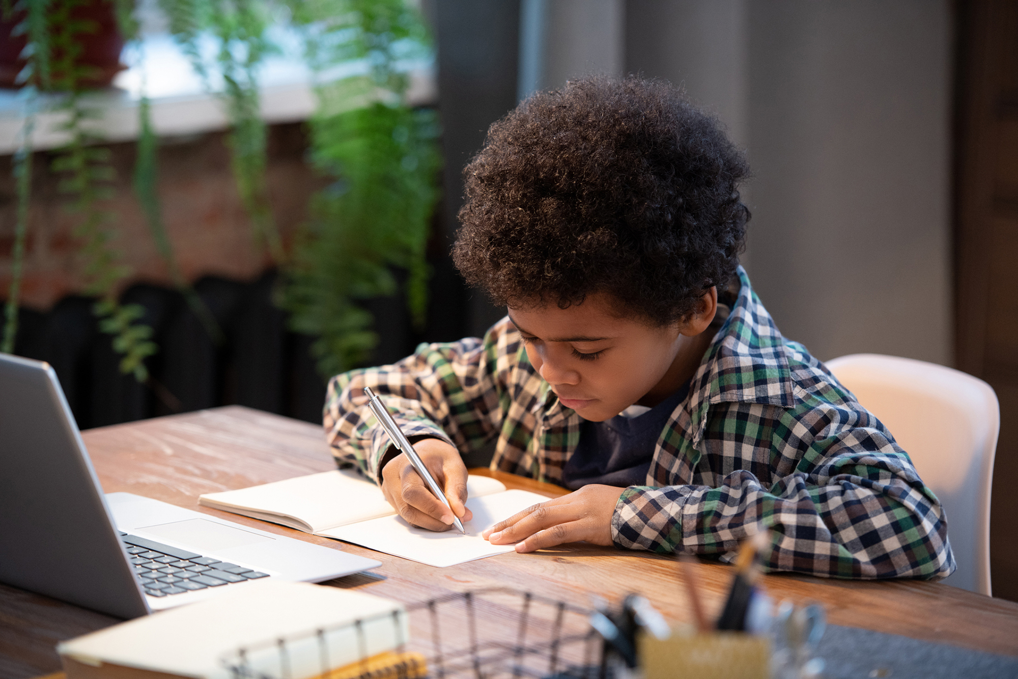 Young child writing in a notebook at a table with a laptop.
