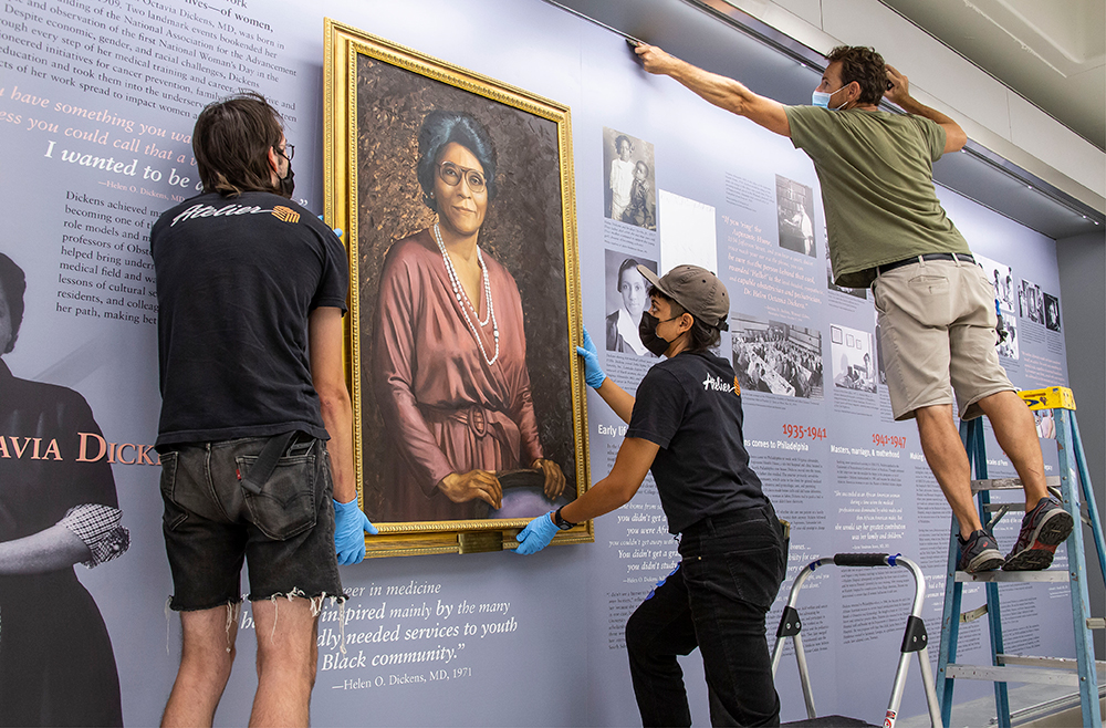 Three masked workers hang a portrait of Helen Octavia Dickens on the wall.