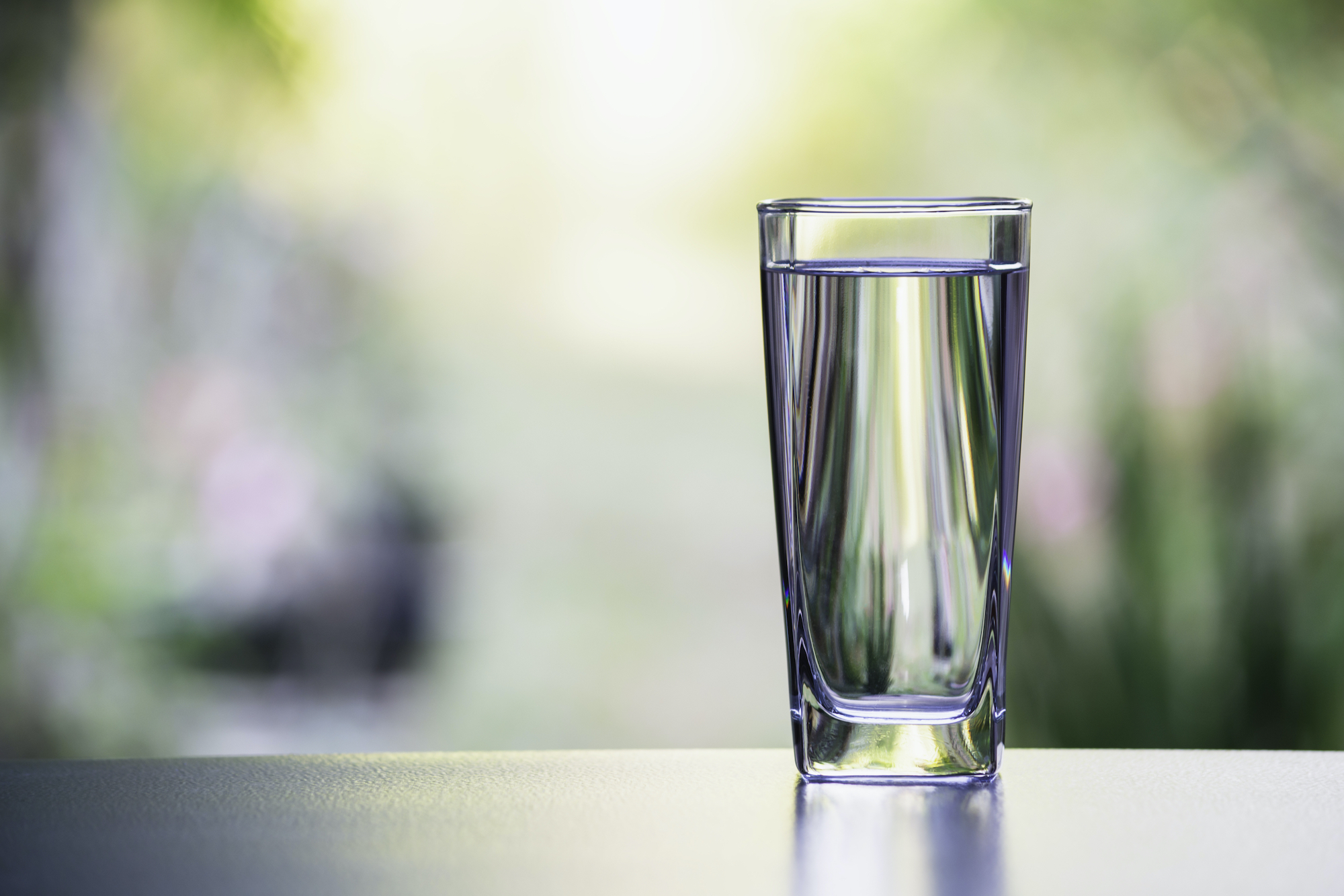 A tall clear glass of water filled almost to the top on a dark-colored countertop. Nature is obvious but blurred in the background.