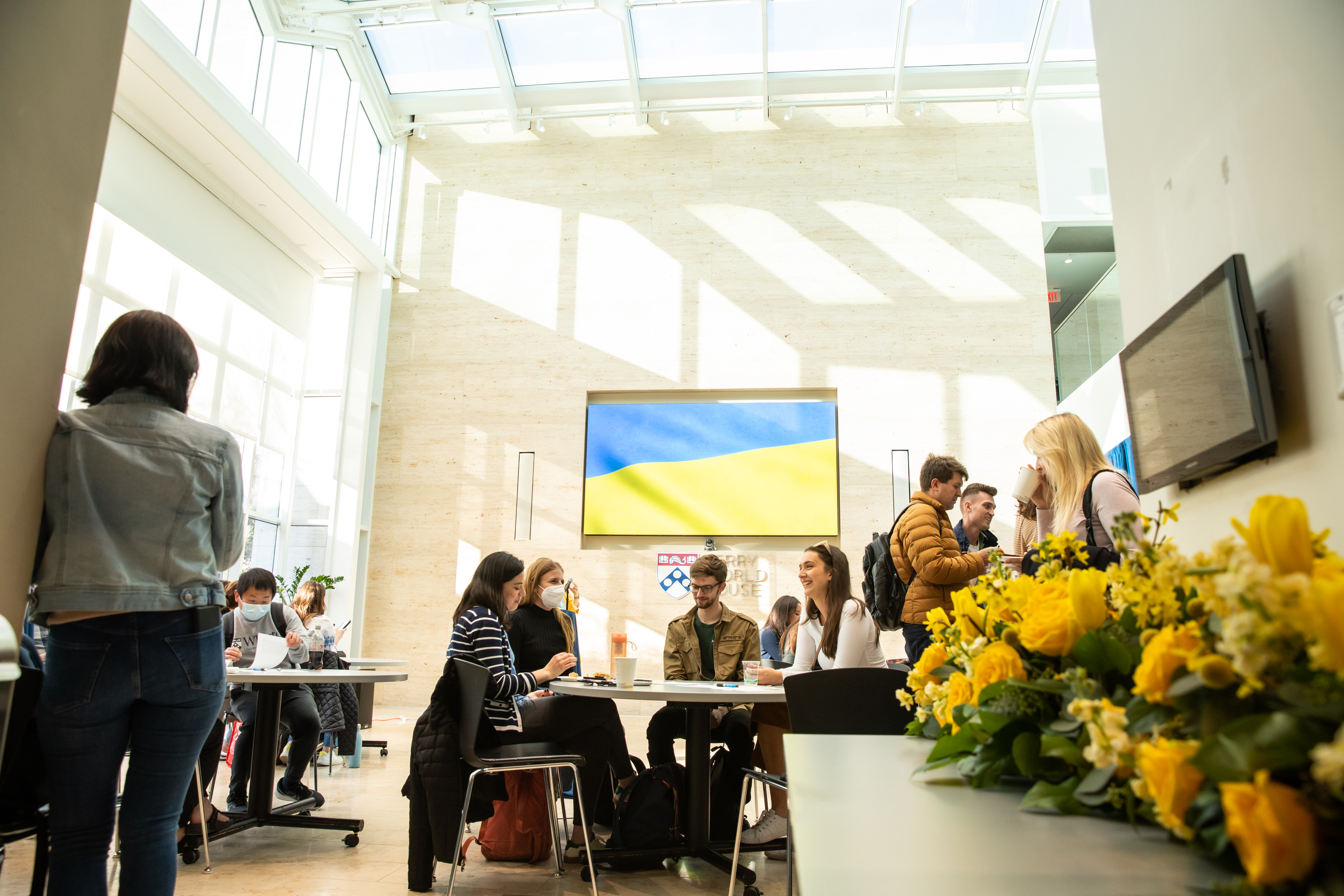 People seated at tables at Perry World House with a digital flag displayed on the back wall and yellow flowers on a table.