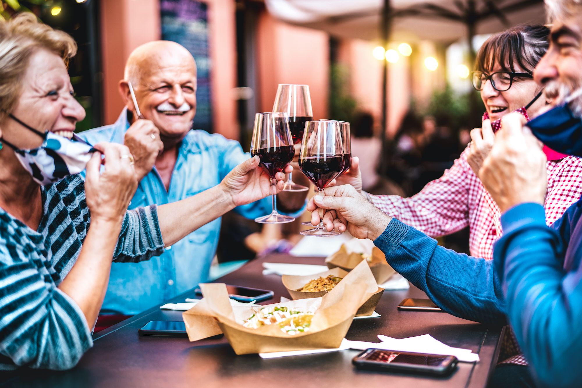 A group of older people at a restaurant clinking half-full wine glasses, with their masks pulled down around their chins to reveal a smile. Food is on the table.