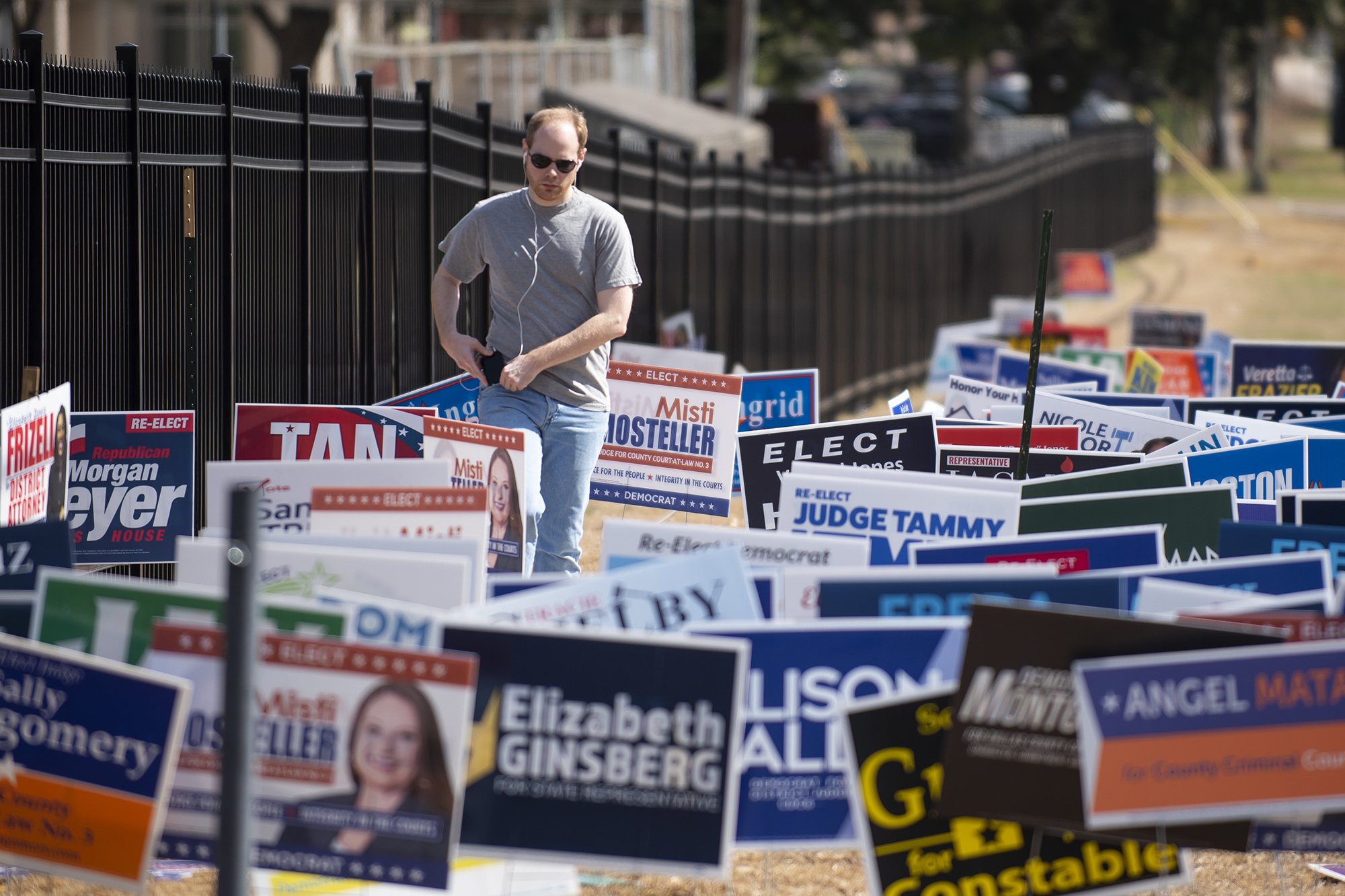 Man in sunglasses with earbuds walks throught a field covered in campaign posters