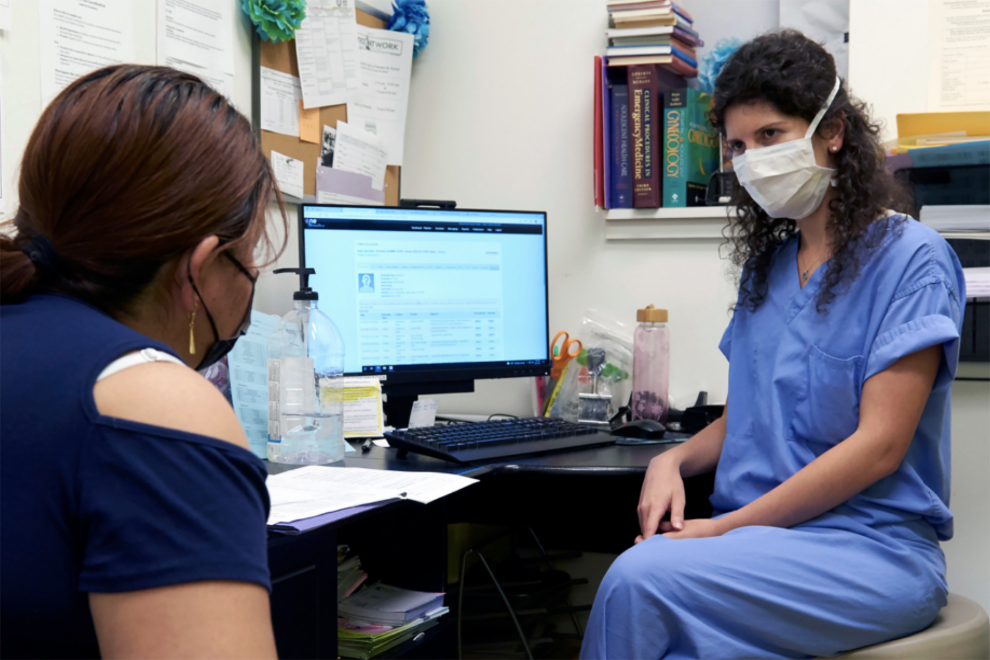 A medical professional sits with a patient at their desk.