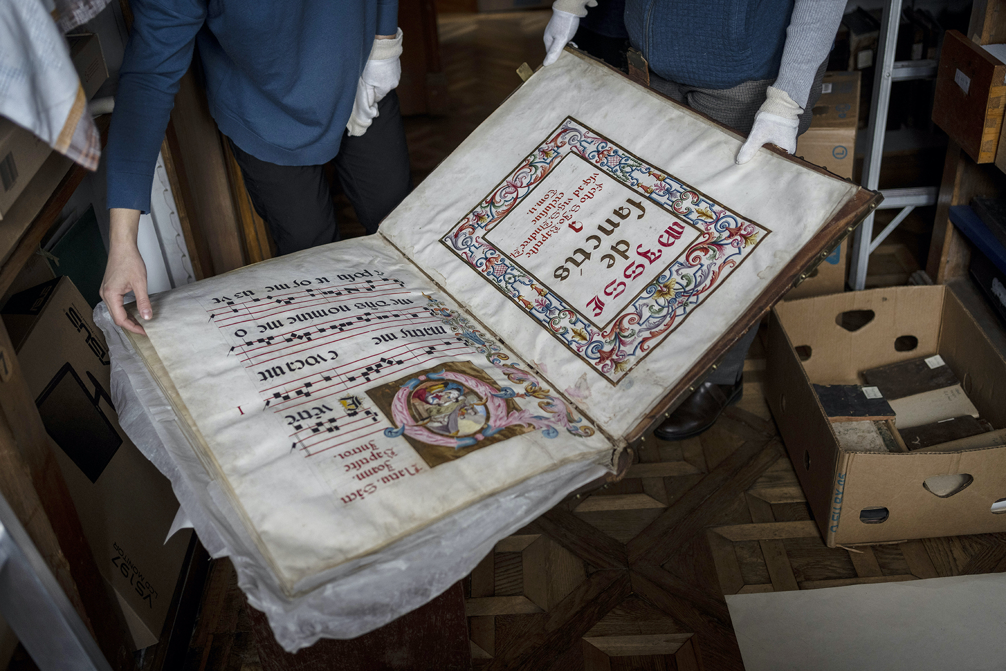 An old and large book upside down on a table, being held by two sets of hands, one gloved, the other not. There are boxes and other materials all around.