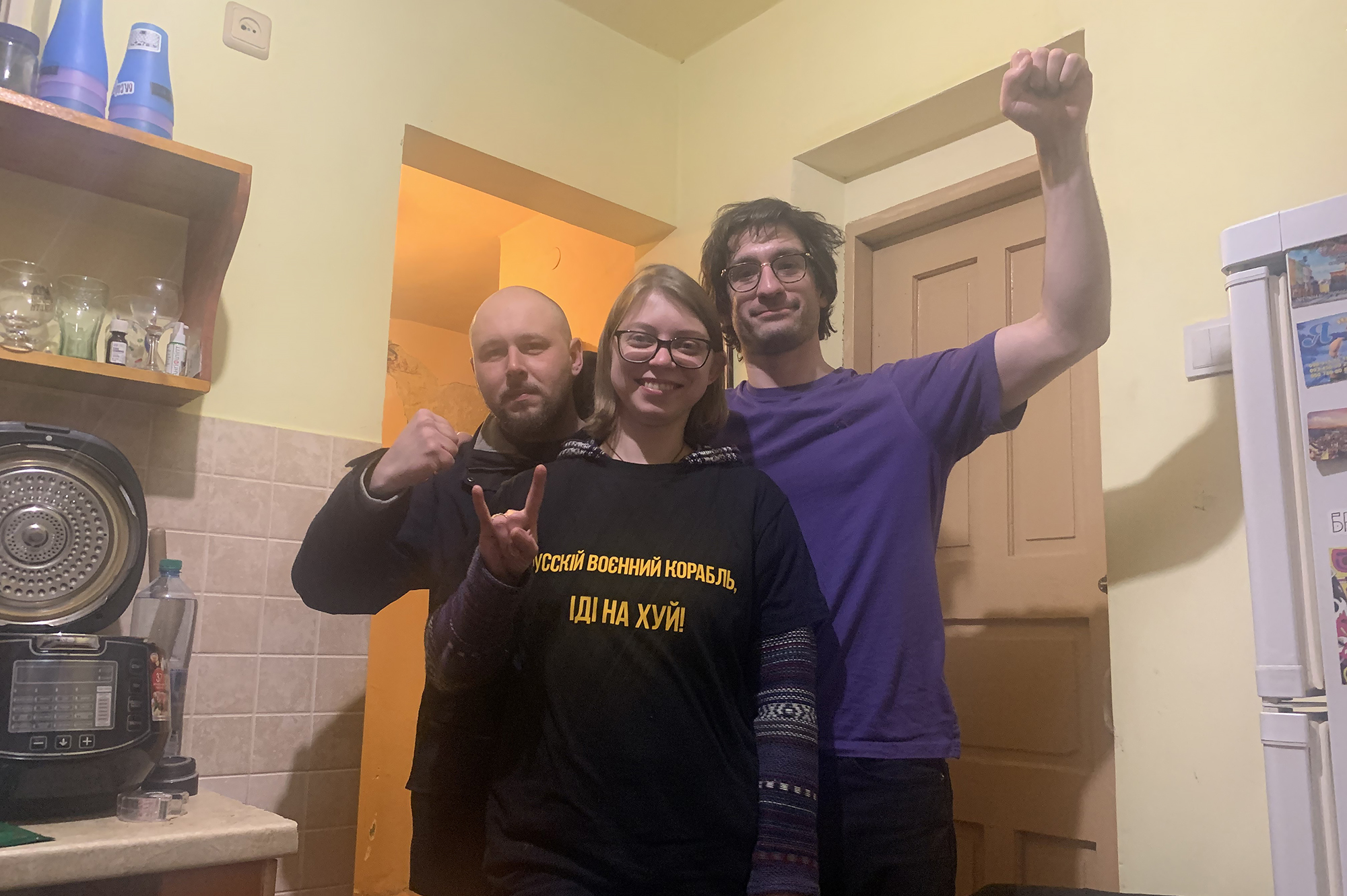 A bald man makes a fist, a woman gives a rock on gesture, and Sam Finkelman shows a raised fist in a kitchen in Ukraine.