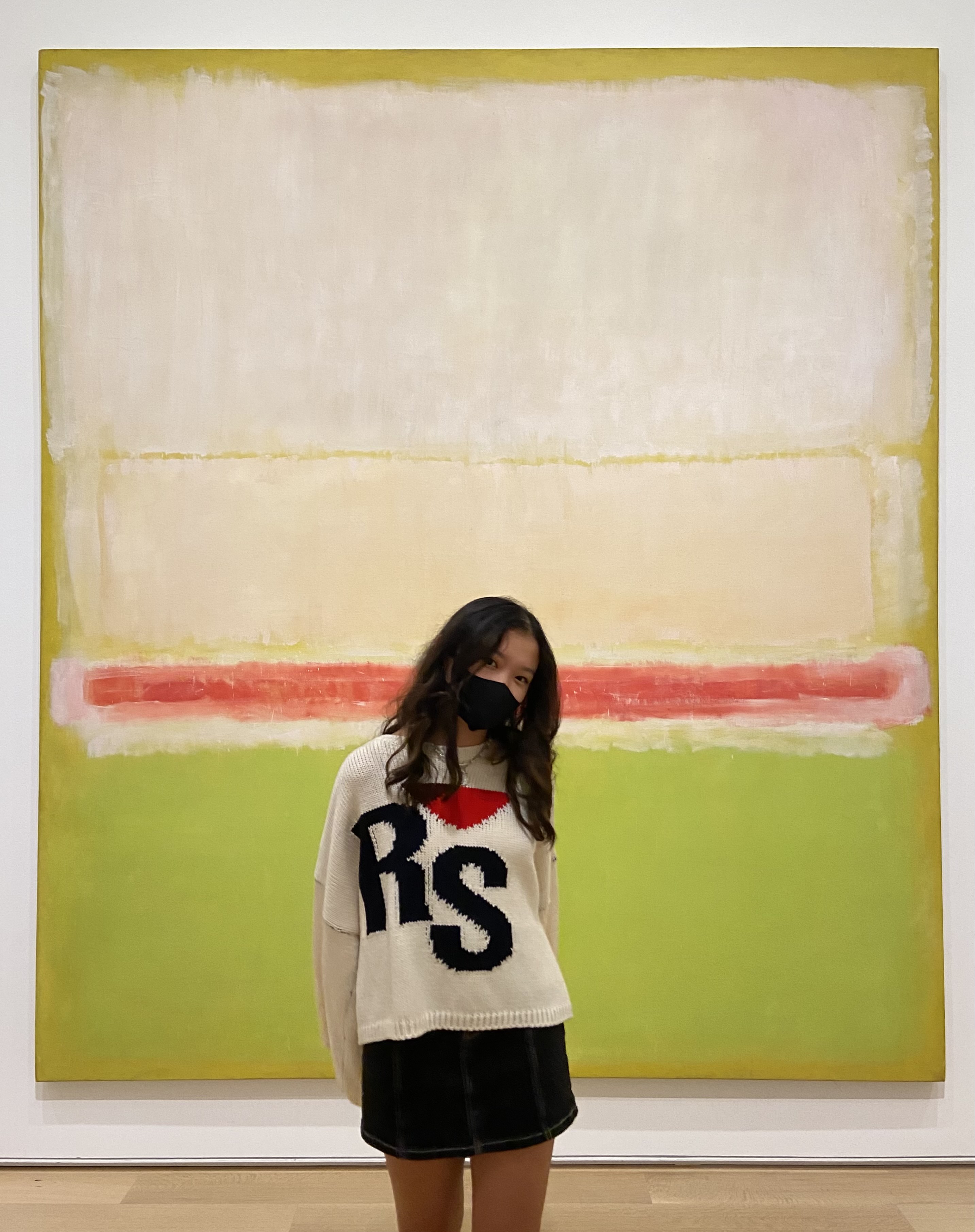Woman wearing a mask and a sweater with the letters "RS" in front of a white, red, and lime green painting in linear blocks of color.
