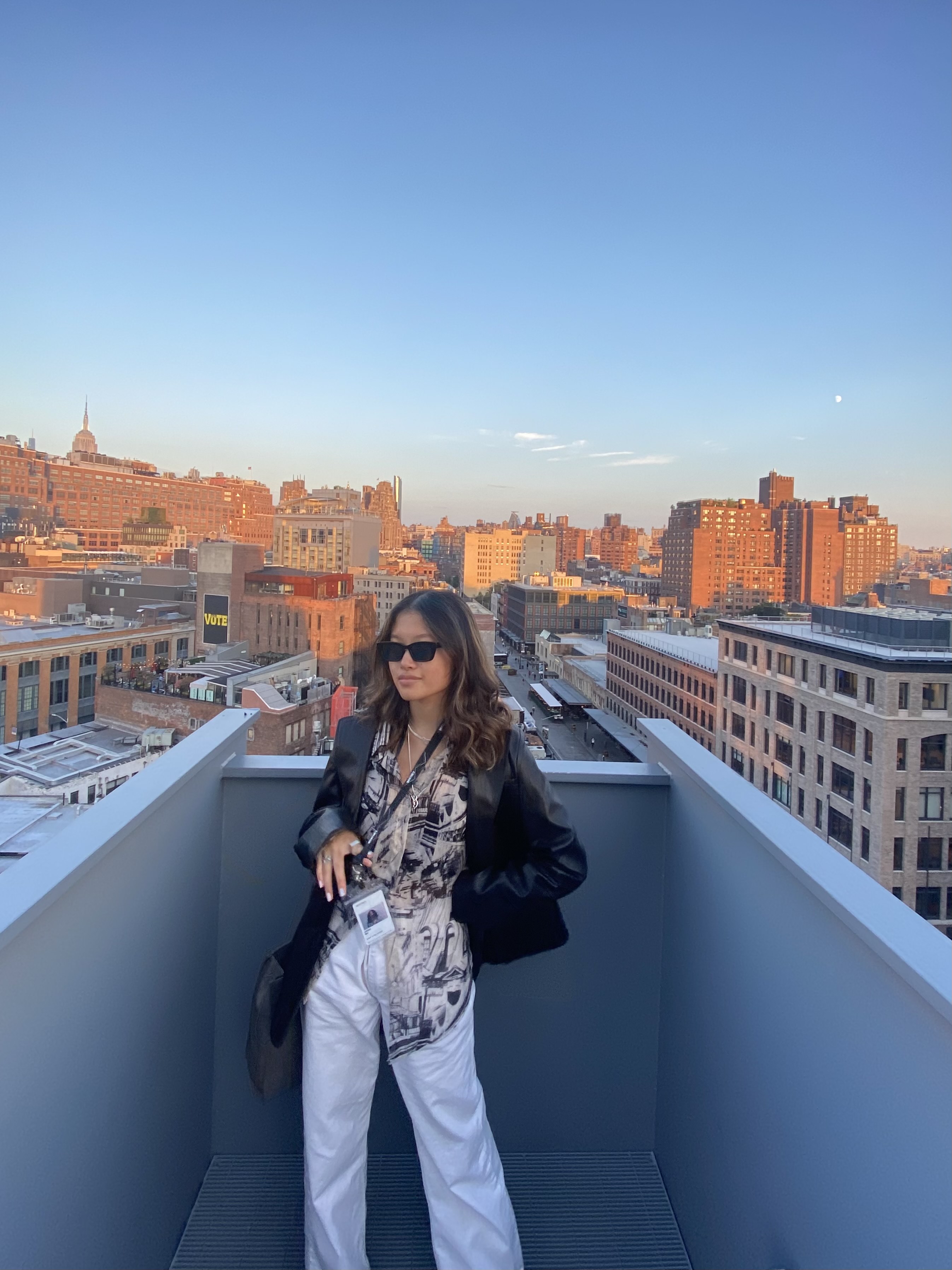 Woman in sunglasses and a leather jacket stands on a rooftop deck with a city skyline behind her.