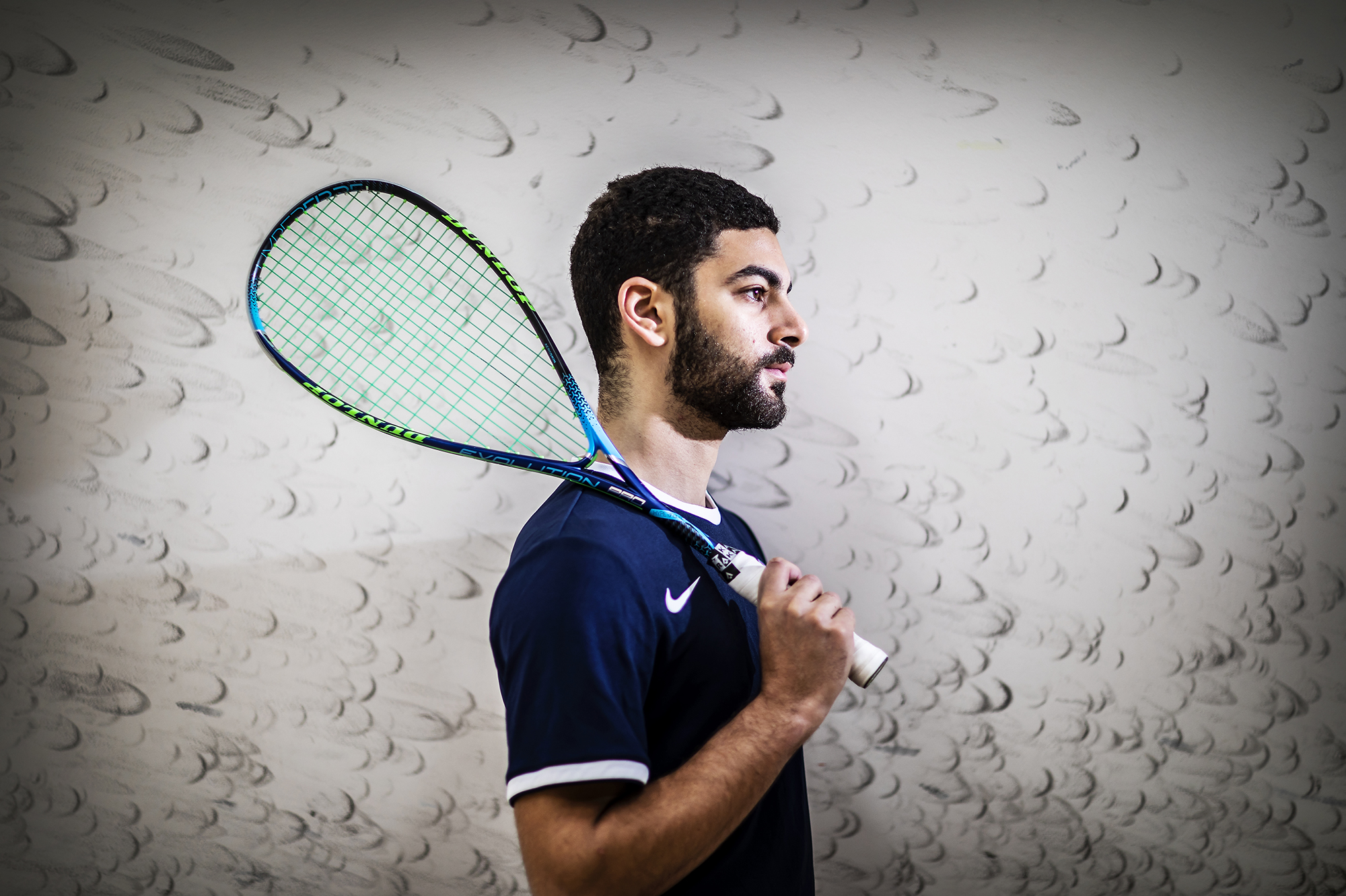 Aly Abou Eleinen holds a racquet on his shoulder in front of a wall showing ball marks.