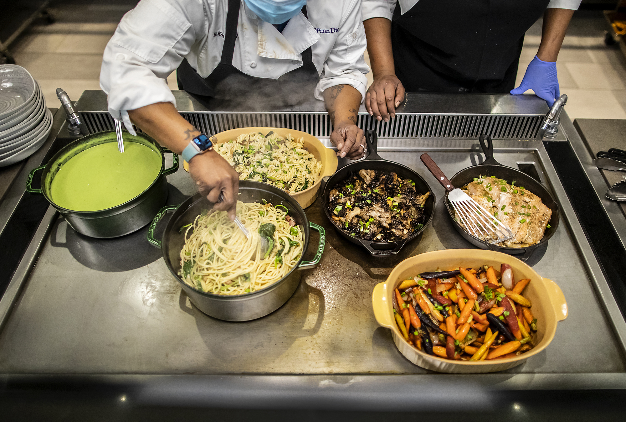 The arms of two people over an industrial-sized kitchen warmer, which holds a pot of green puree, two pans of pasta, a cast iron skillet of mushrooms, another cast iron skillet of fish with a spatula on top, and a bowl of multi-colored carrots. A stack of bowls sits off to the left of the image.