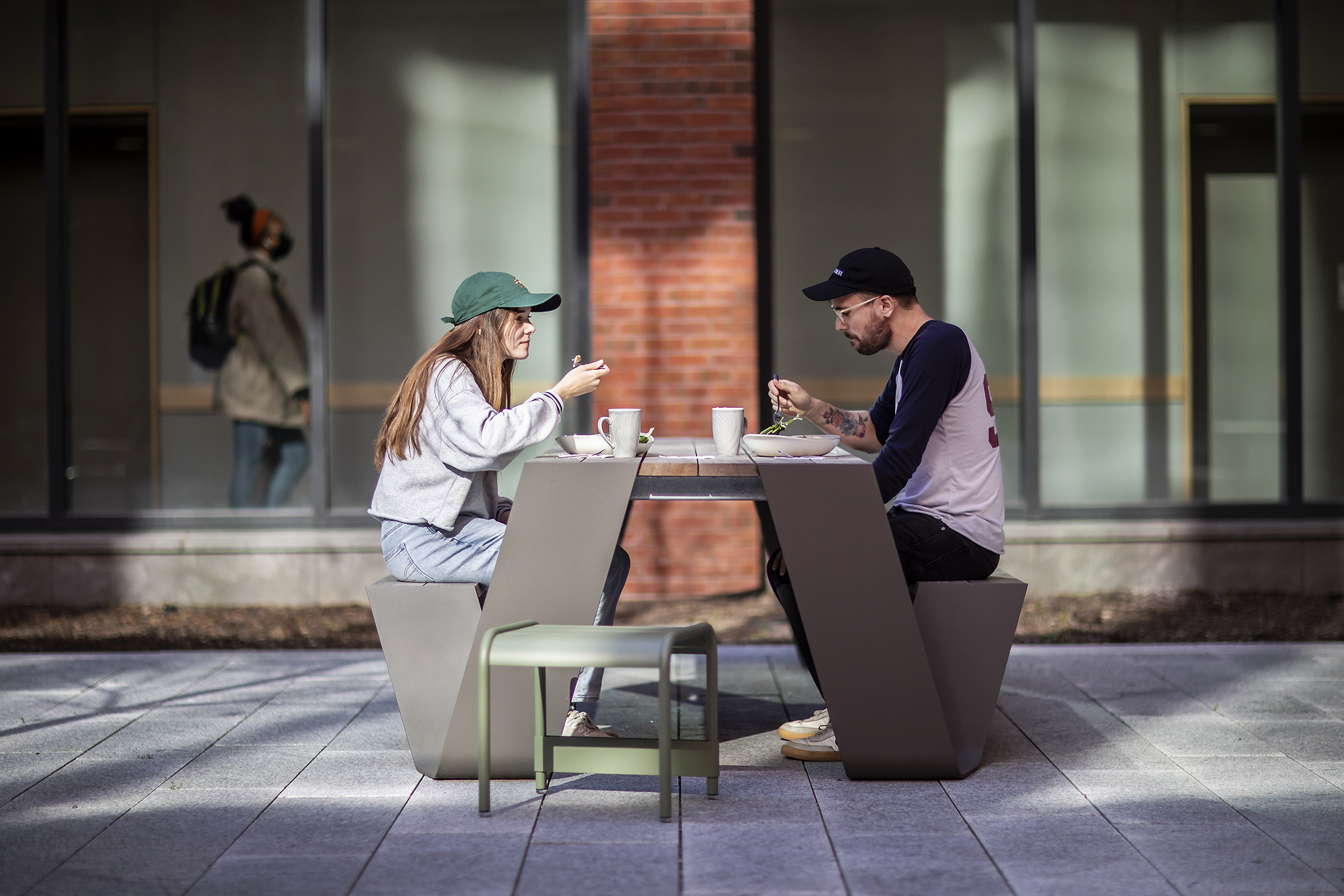 Two people sitting outside at a table eating food, with a person inside blurred in the background.