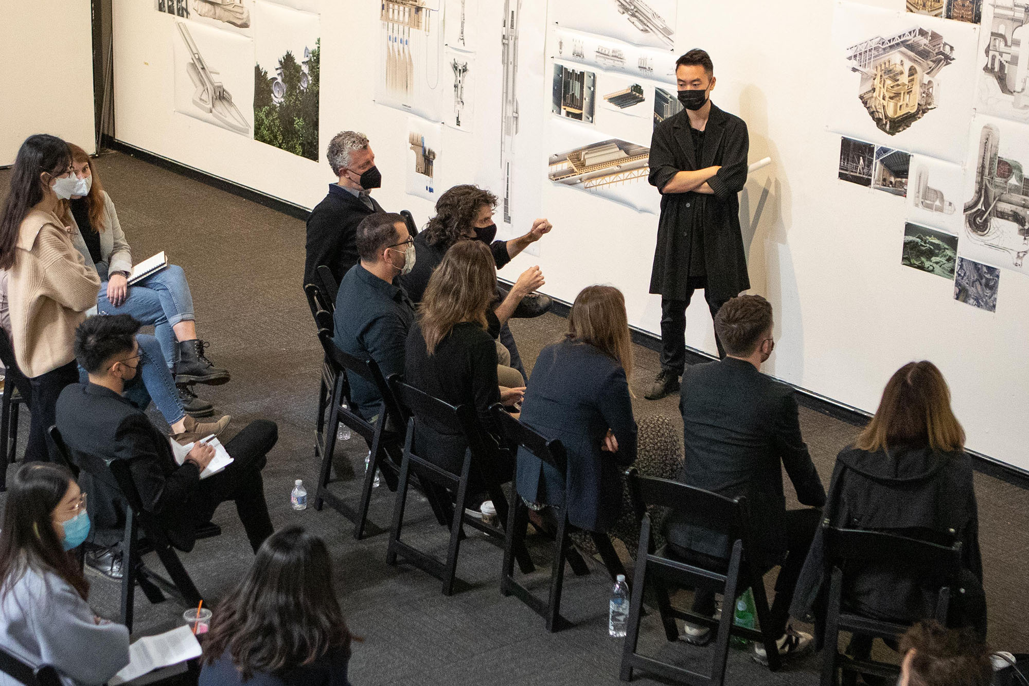 A person stands in front of a wall of photos and architectural renderings before a group of seated people.