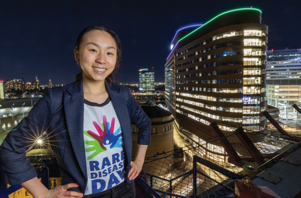 Magnolia Wang stands on an outdoor viewing point with HUP in the background illuminated in green and purple lights.