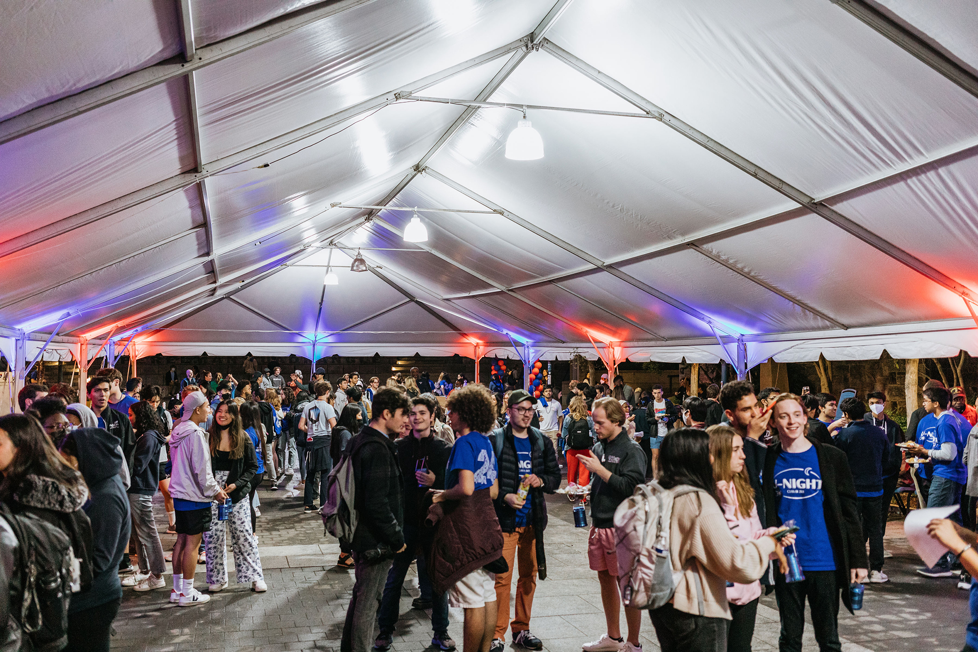 A large group of Penn students gather at night under a large white tent.