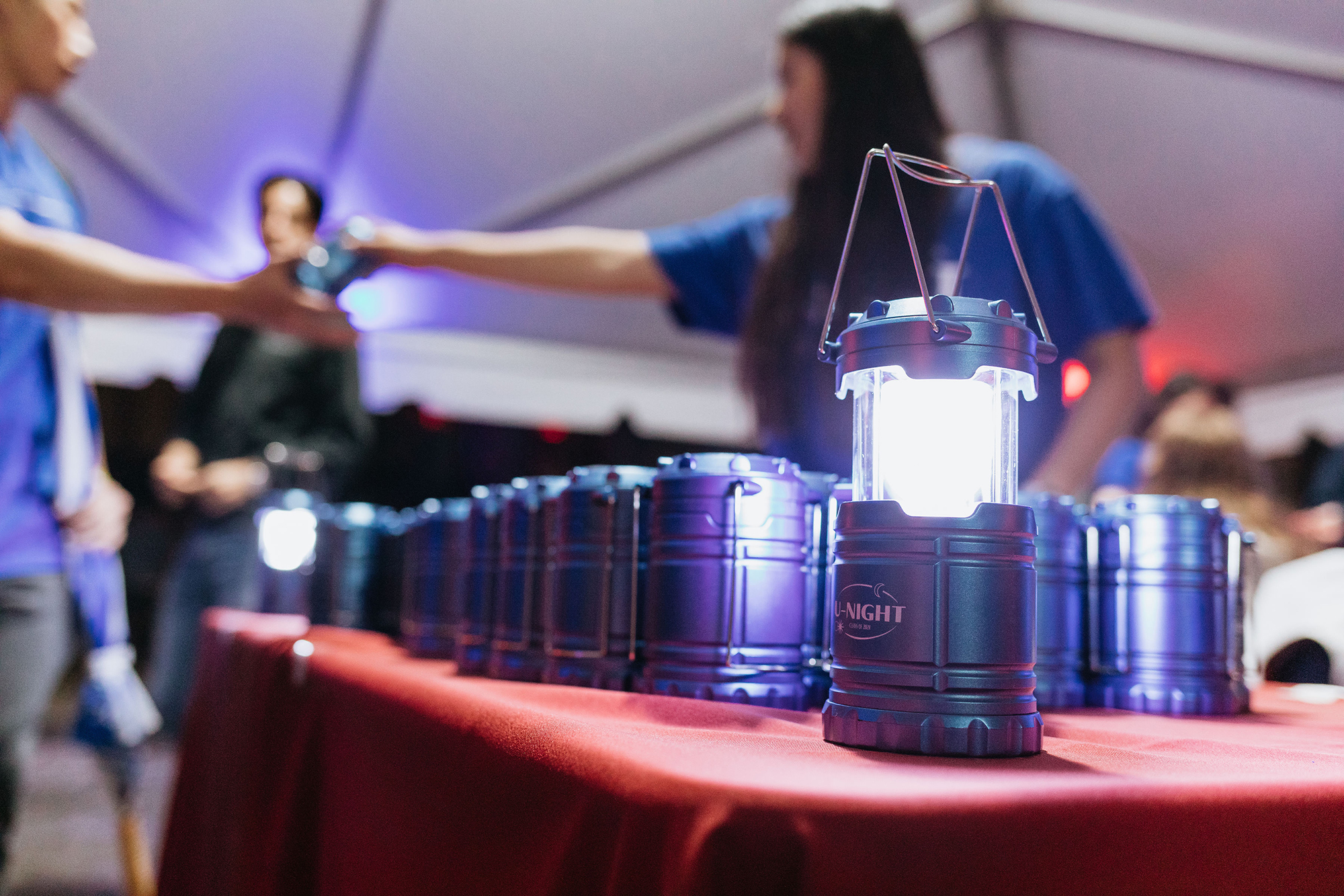 An illuminated battery-powered lantern is lit on a table among other lanterns, behind it a person hands a student a lantern.