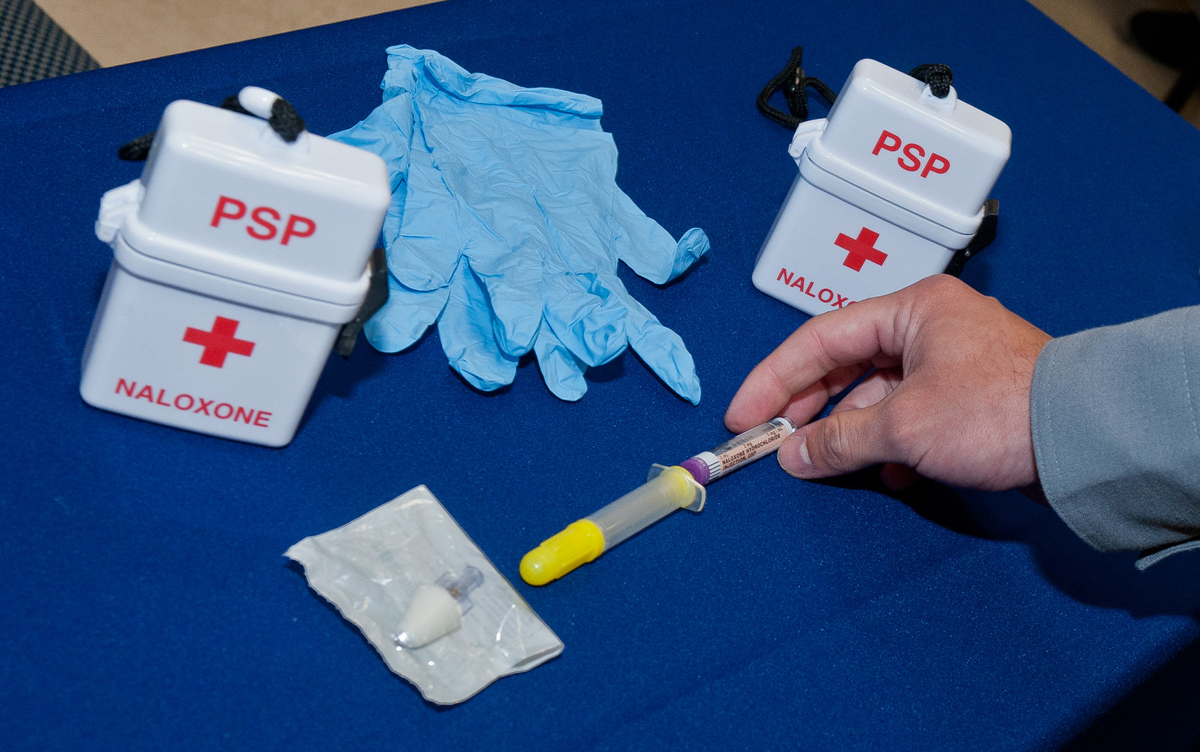 A hand holding a syringe on a table with latex gloves and two naloxone kits.
