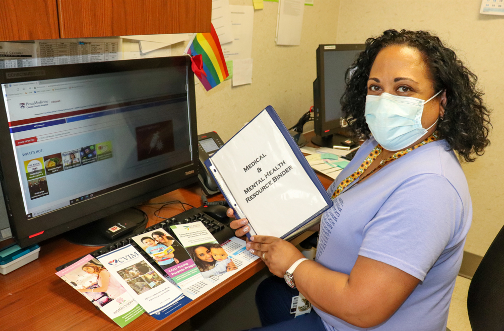 Lissette “Mitzy” Liriano sits at a desk with a computer, wearing a face mask, holding a binder that reads “Medical and mental health resource binder.”