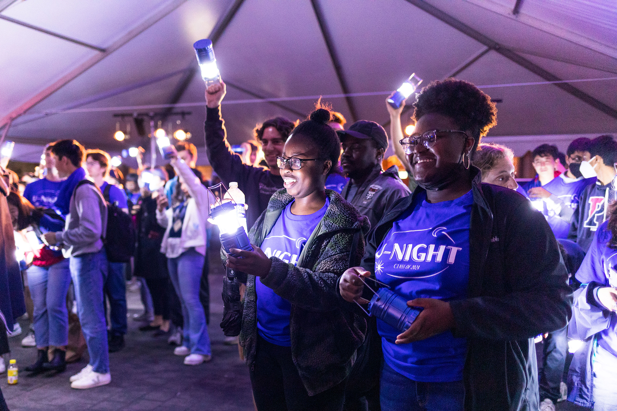 A group of Penn undergrads wearing U-Night shirts hold lanterns under a large party tent.
