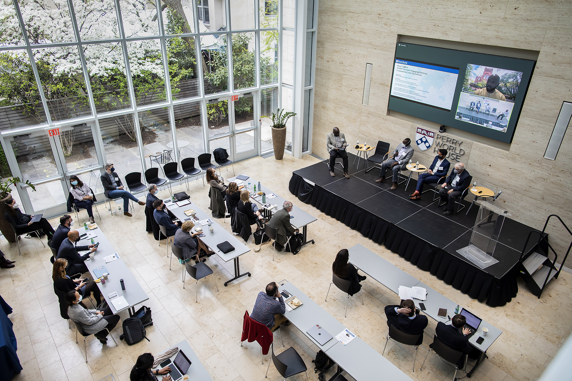 Panelists sit on a stage at Perry World House, while another is on a Zoom screen behind them