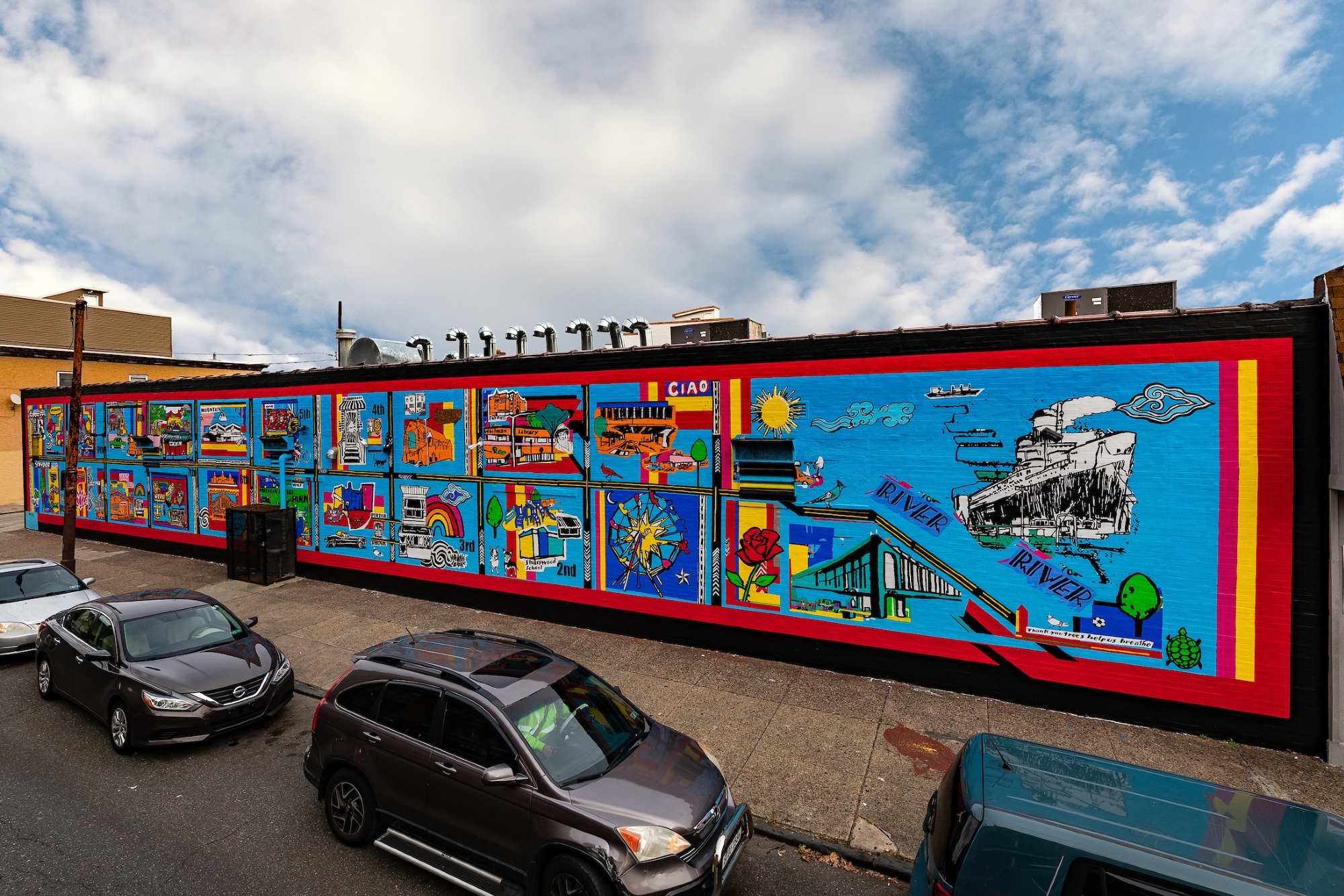 mural with many illustrations in bright colors on a long wall with a sidewalk and cars