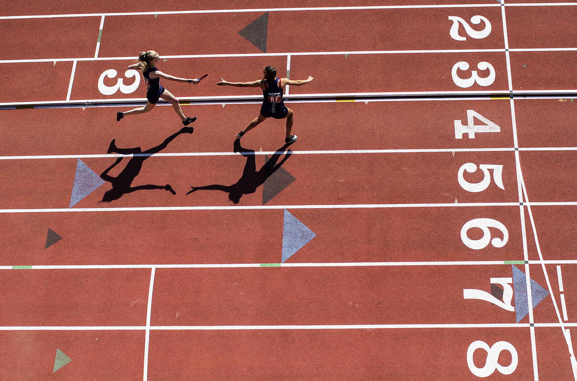 Overhead view of one runner on a track passing a baton to another.