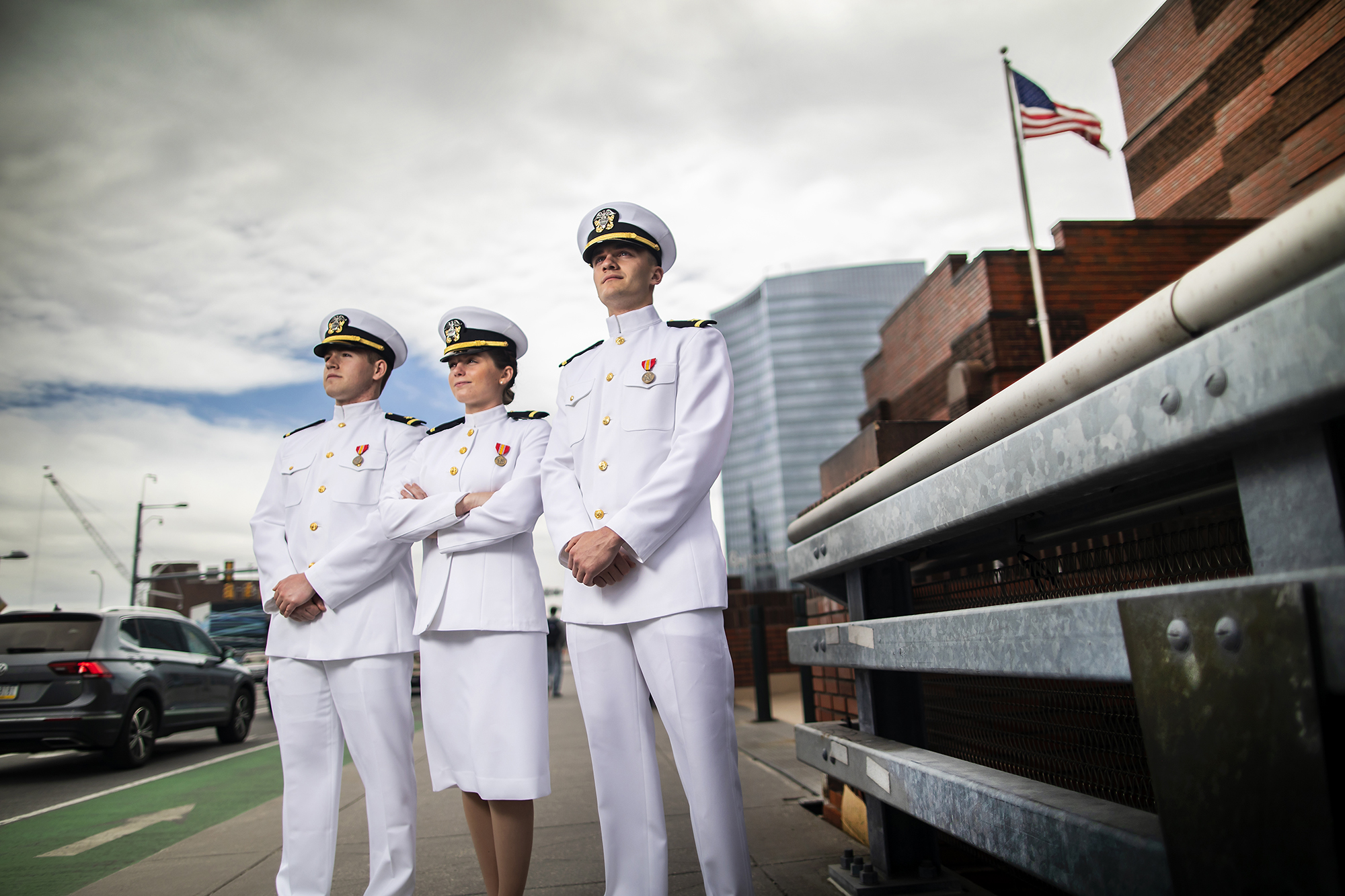 Three students in dress whites stare into the distance with a busy bridge on their left and the American flag in the background. 