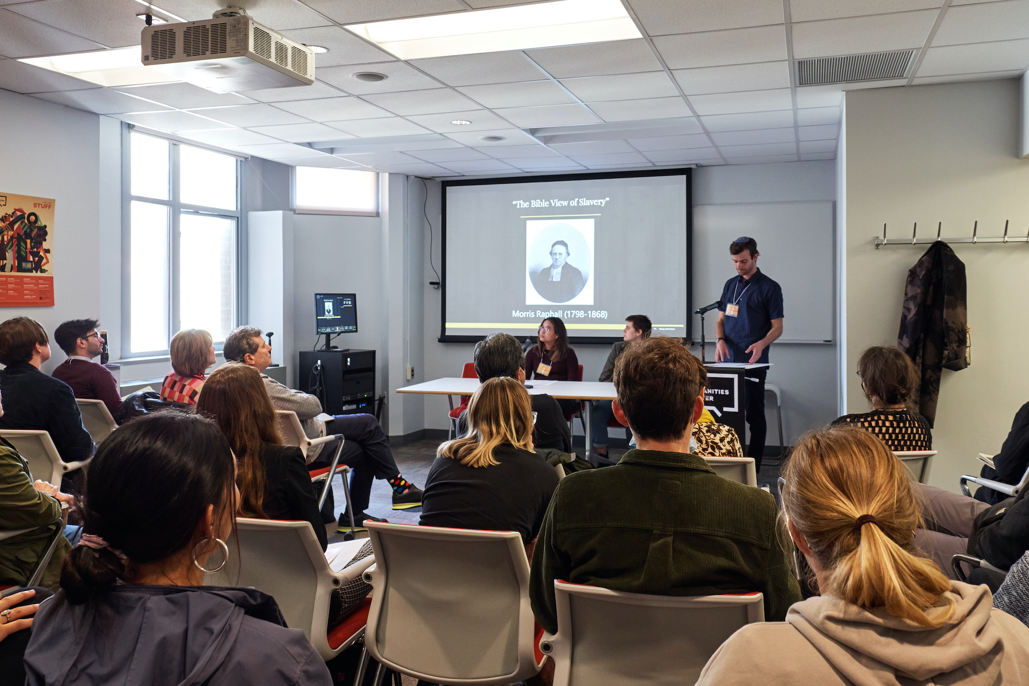 A group of students and professors listen to Samuel Strickberger present his research. The screen reads "The Bible View of Slavery"