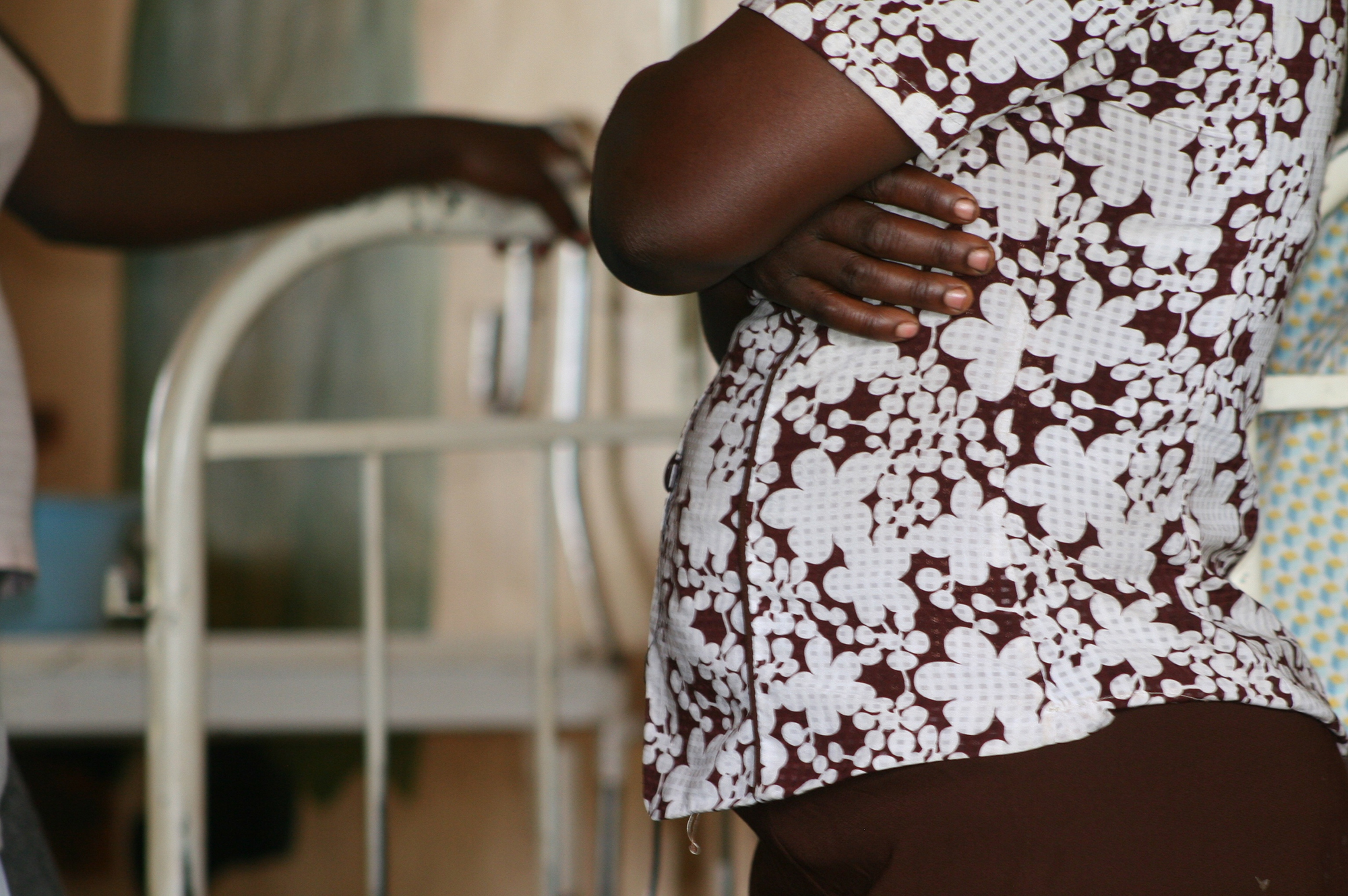 Torso of a pregnant person with arms crossed standing by a hospital bed in Botswana.