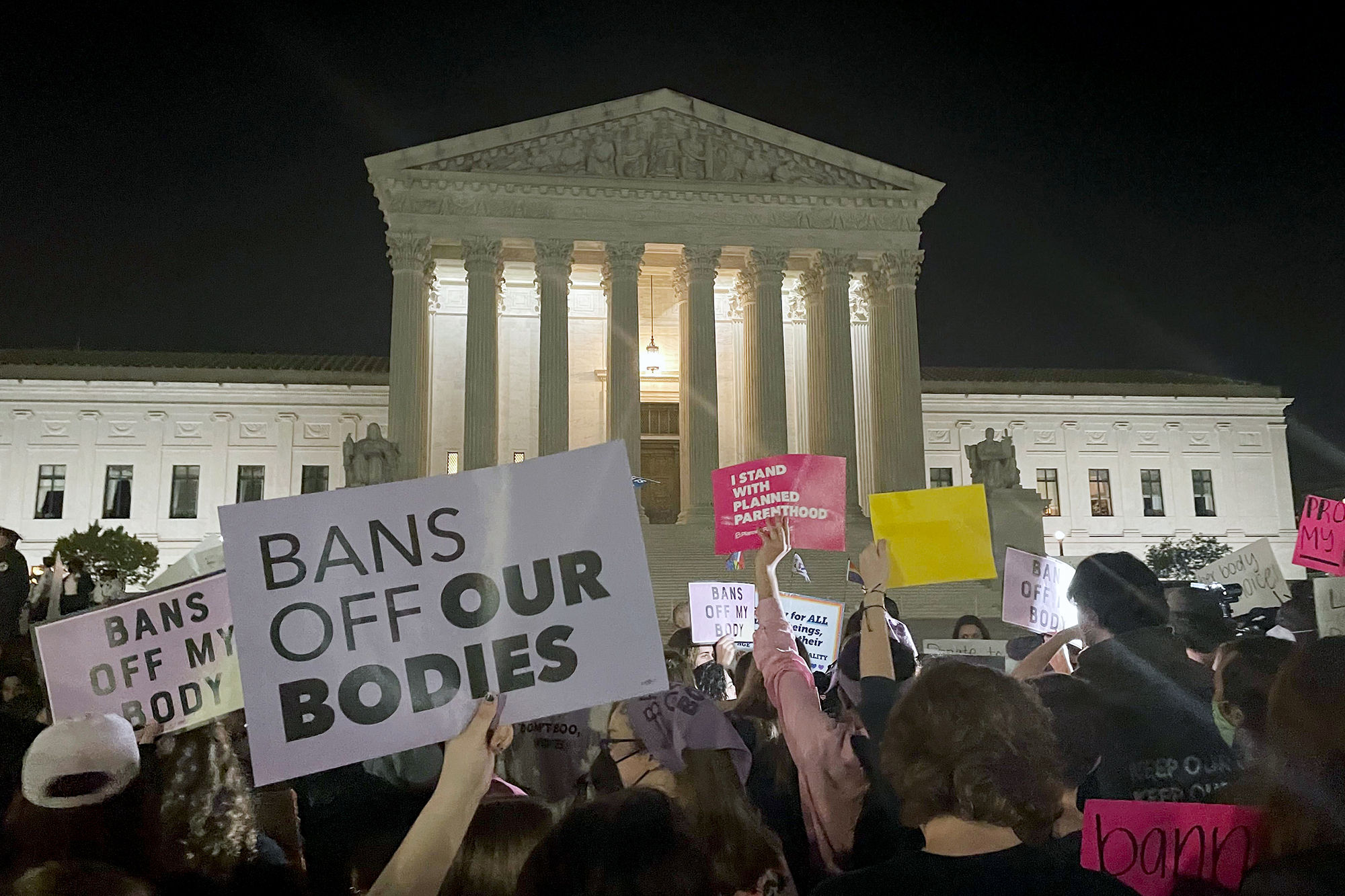 Crowd in front of U.S. Supreme Court at night holding pro-choice signs like BANS OFF OUR BODIES and I STAND WITH PLANNED PARENTHOOD.