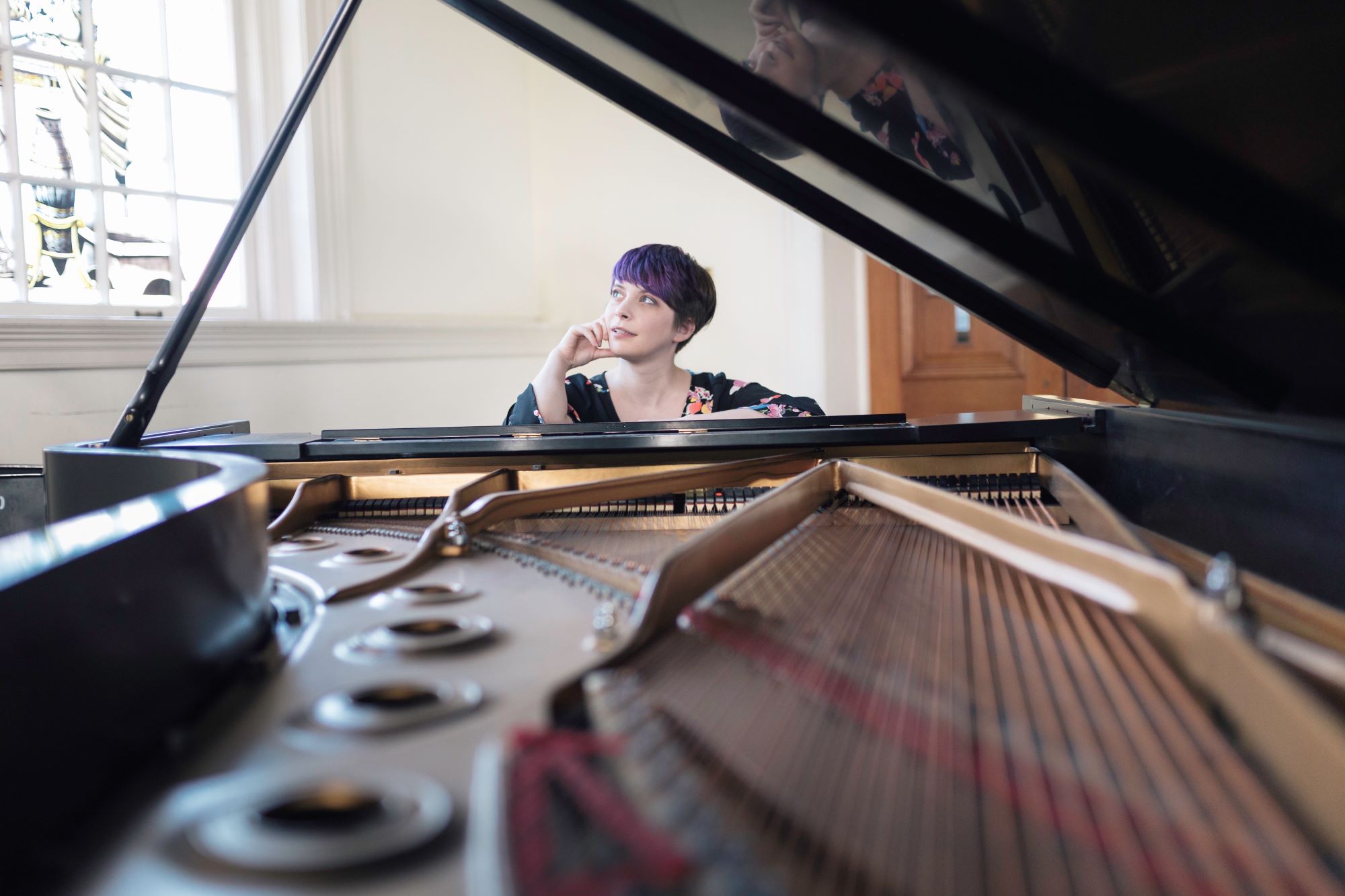 A woman with purple hair sits at a piano and stares off into the distance.