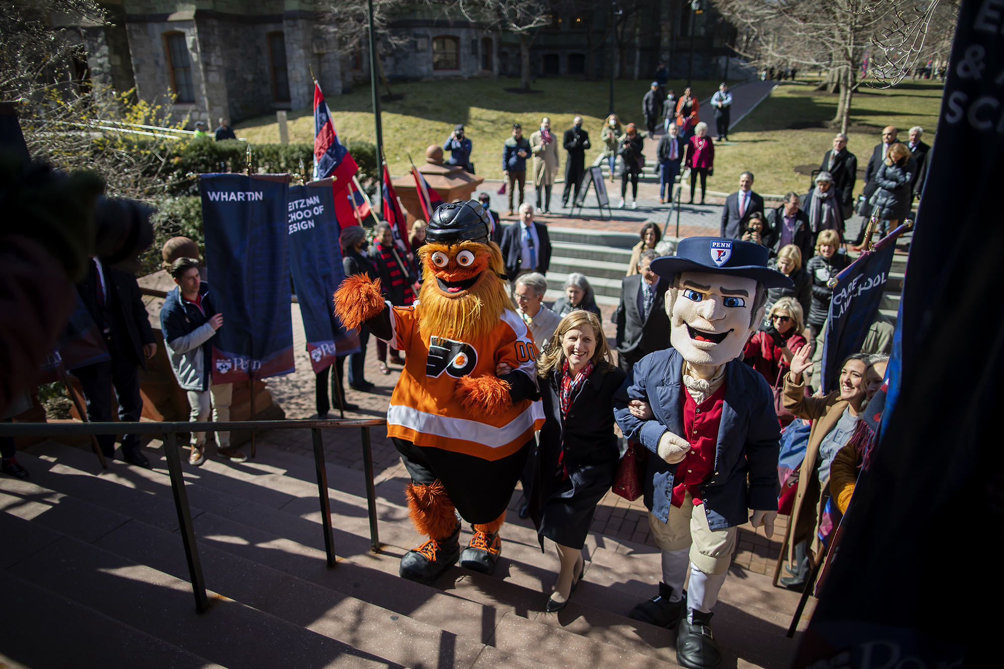 Liz Magill walks up the steps of Fisher Fine Arts Library linking arms with Gritty and the Penn Quaker mascot.