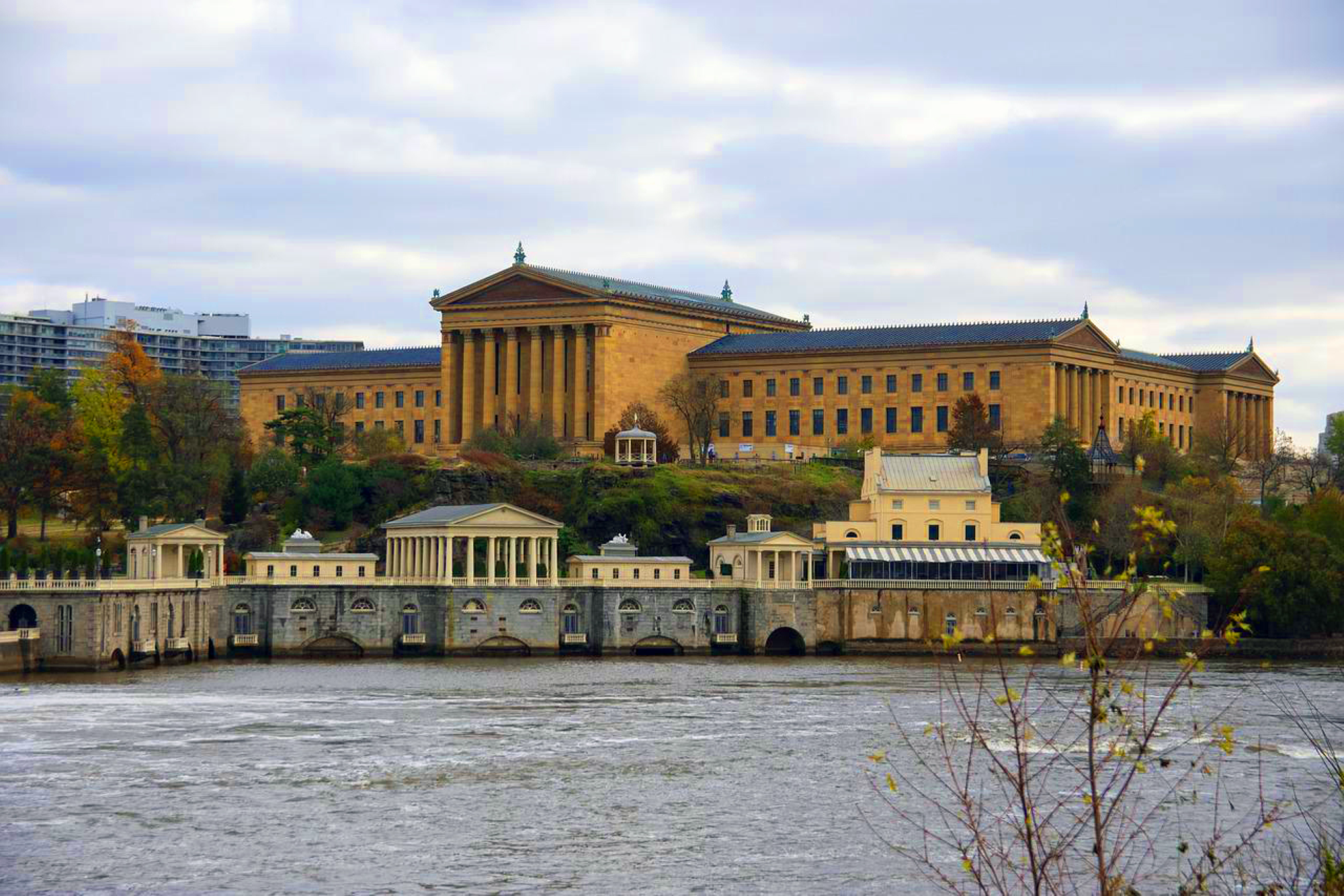 A view of the Philadelphia Museum of Art, a long building of orange brick and blue angled roofs. The Schuylkill River flows in the foreground.