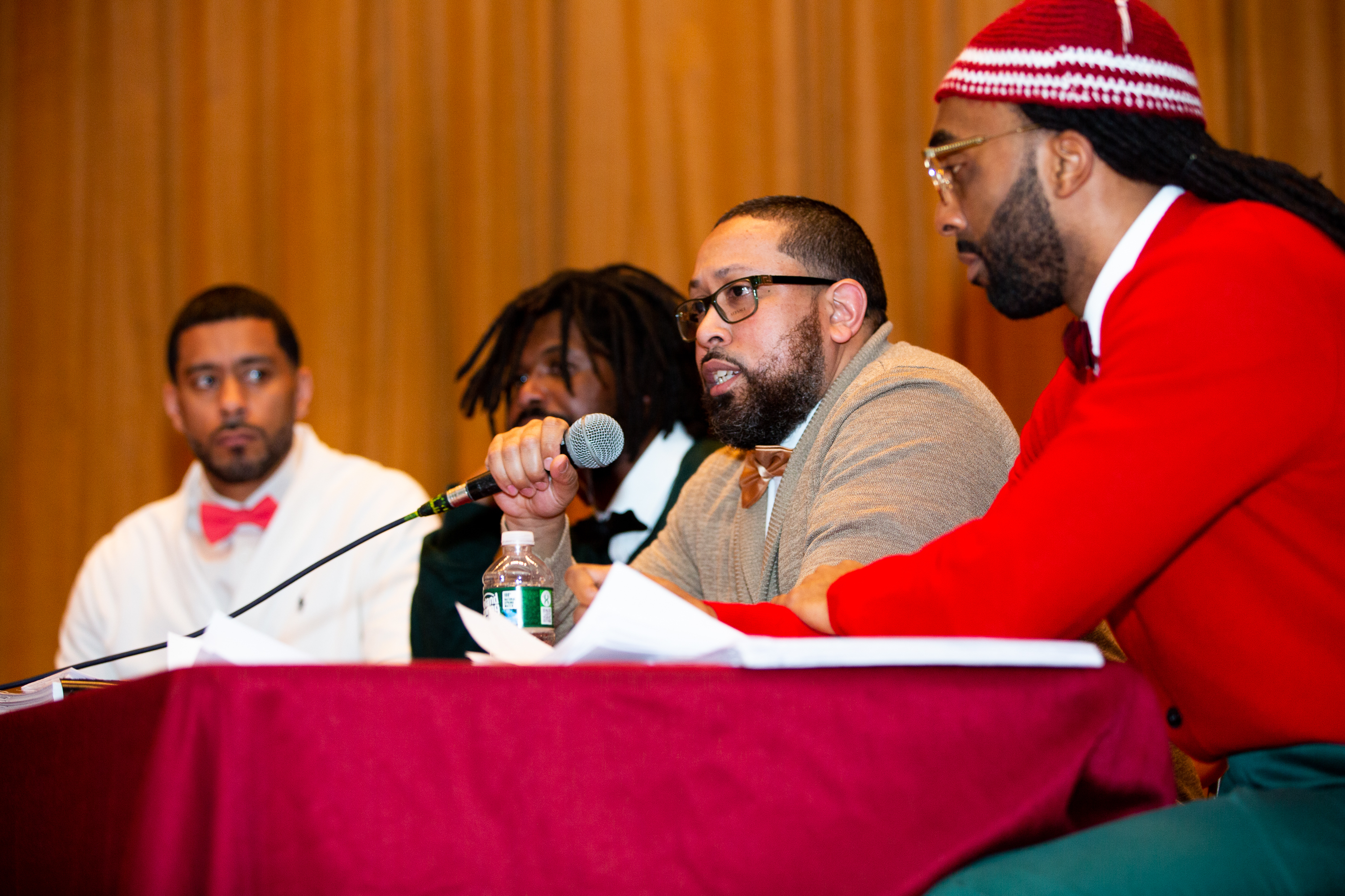 Four members of the Bard Prison Initiative Debate team sit at a table, while one speaks into a microphone