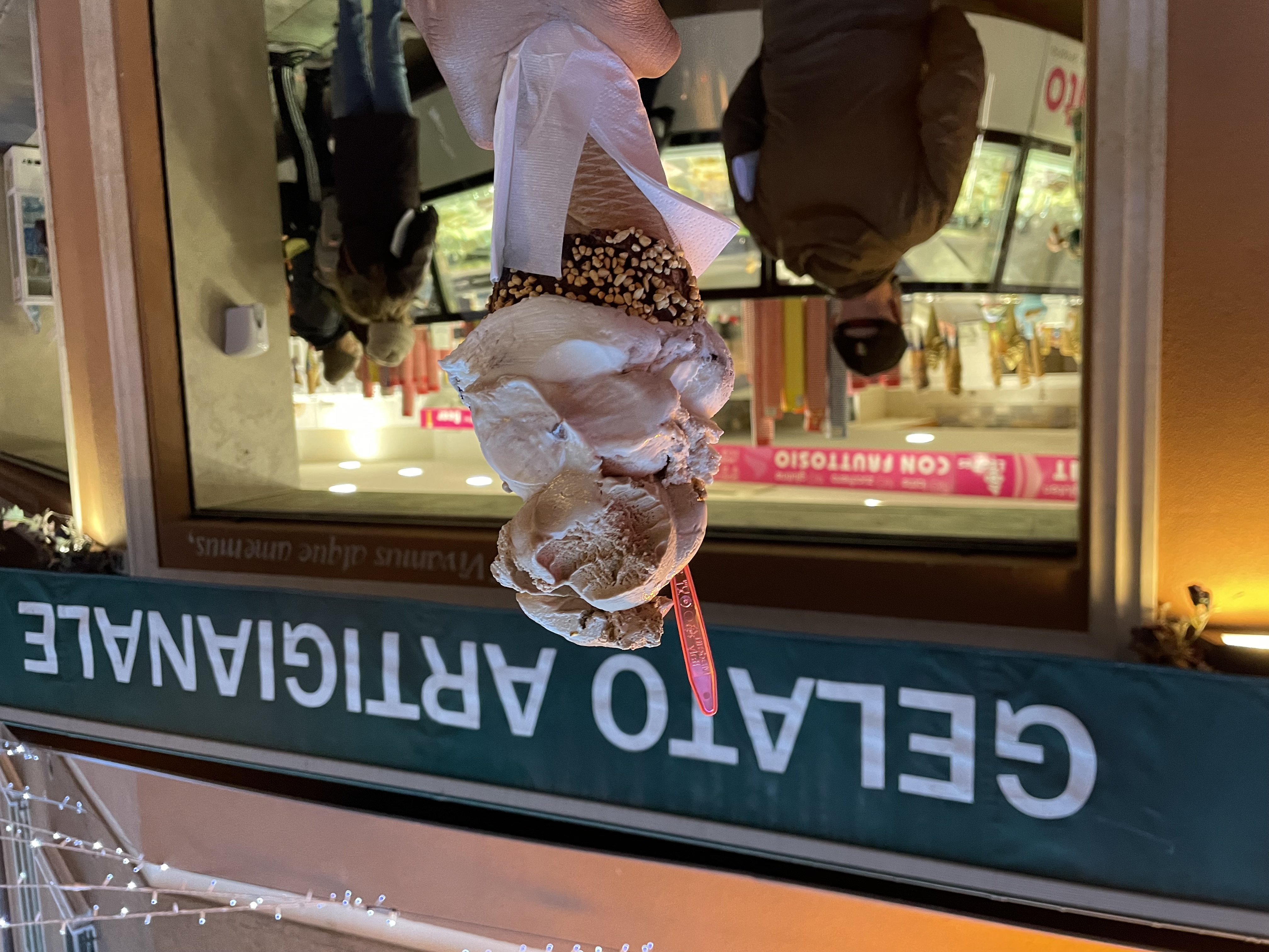 An image of a double-decker ice cream cone with a bite taken out. The sign in the background reads "Gelato Artigianale"