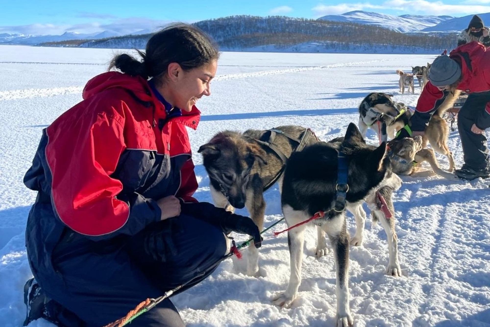 Kiersten Thomas crouching in snow with dogs attached to dogsled on frozen tundra with mountains in the background